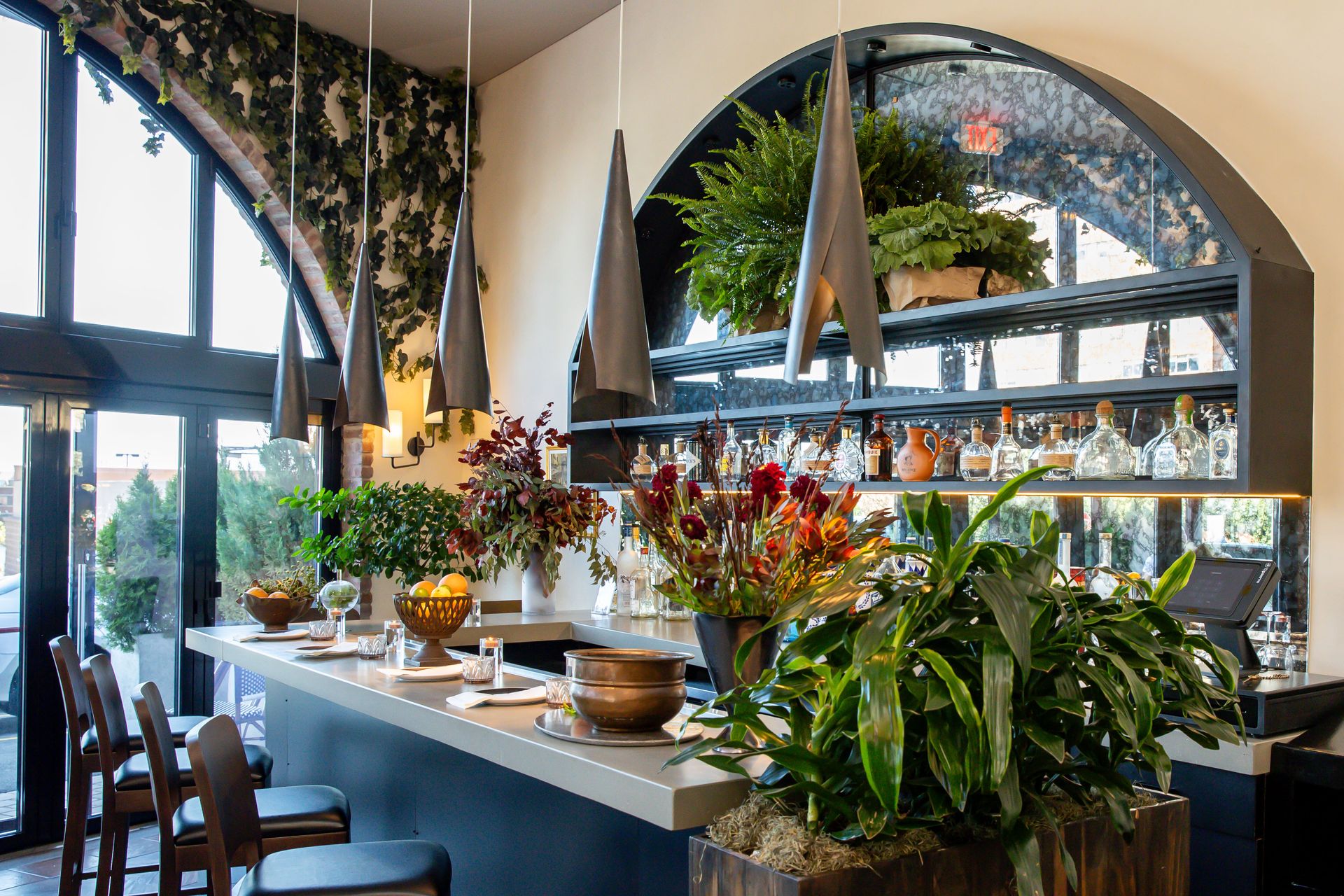 Bar interior with plants, hanging lights, mirrored shelves, and an arched window.