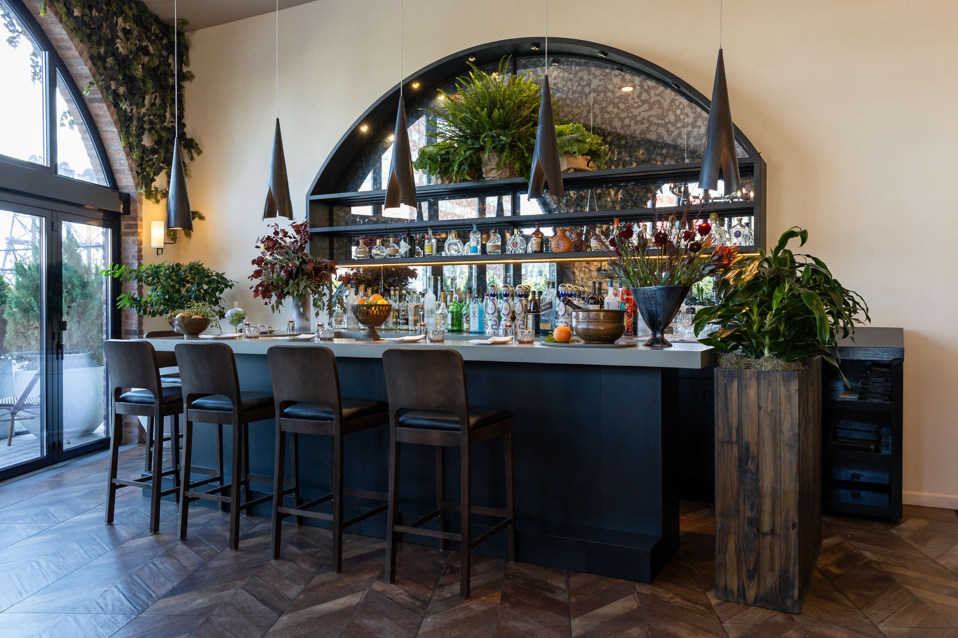 Bar with stools, shelves of bottles, and plants, under an arched mirror, in a room with a tiled floor.
