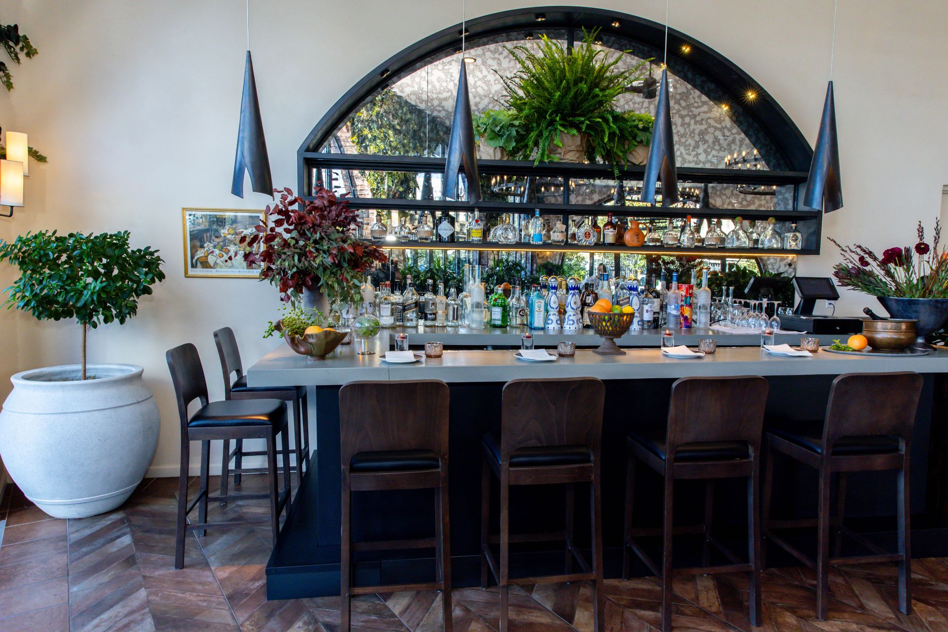 Bar with stools, glassware, bottles, and an arched window backdrop featuring plants.