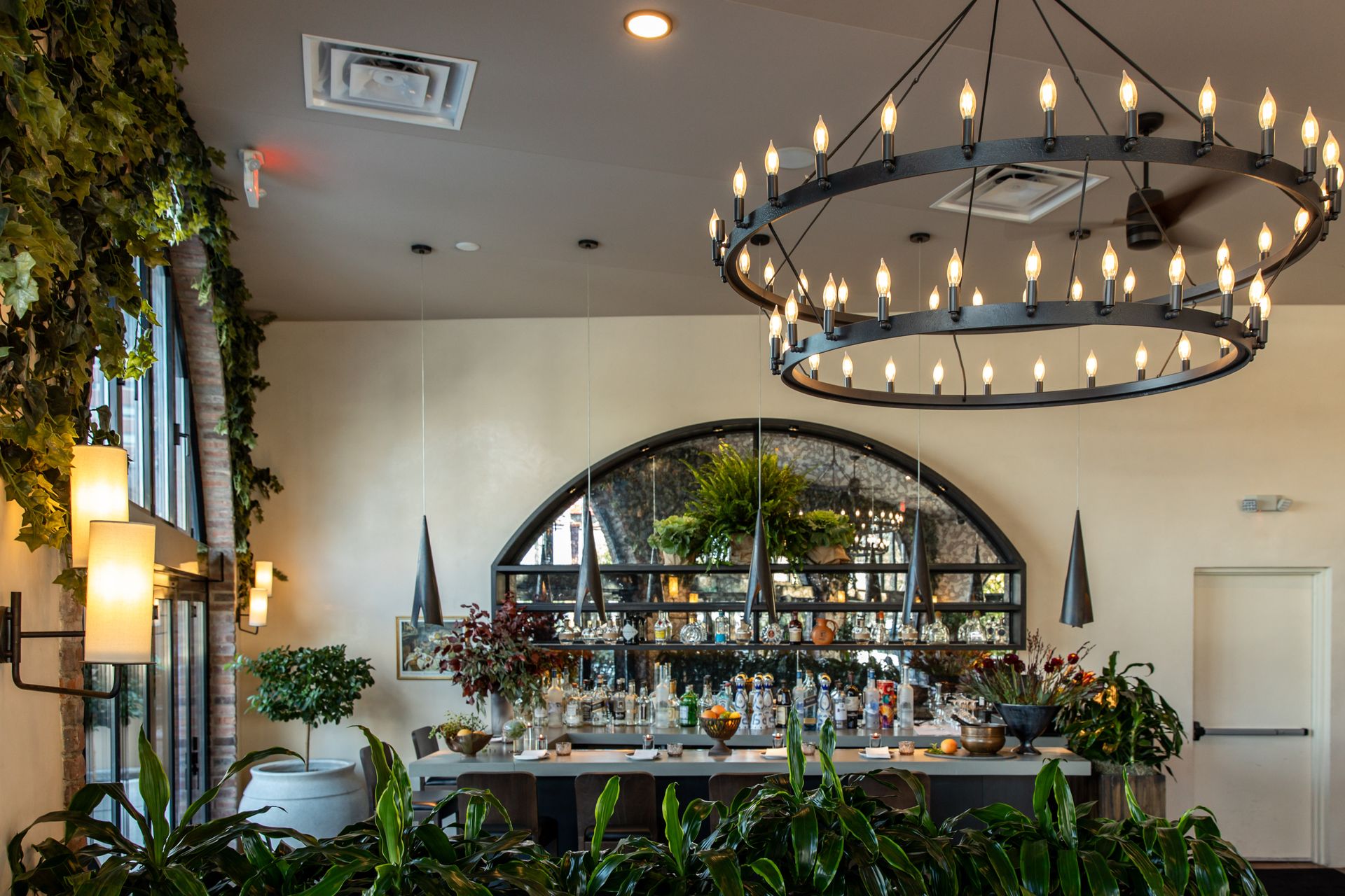 Bar interior with a large circular chandelier, plants, and an arched mirror.