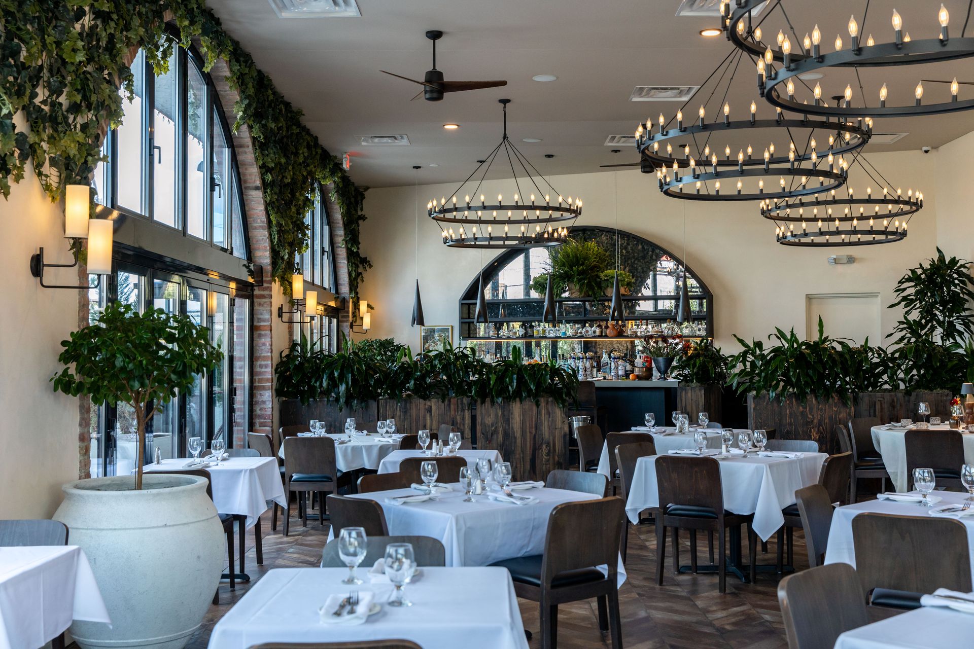 Elegant restaurant interior with white tablecloths, round chandeliers, and greenery.