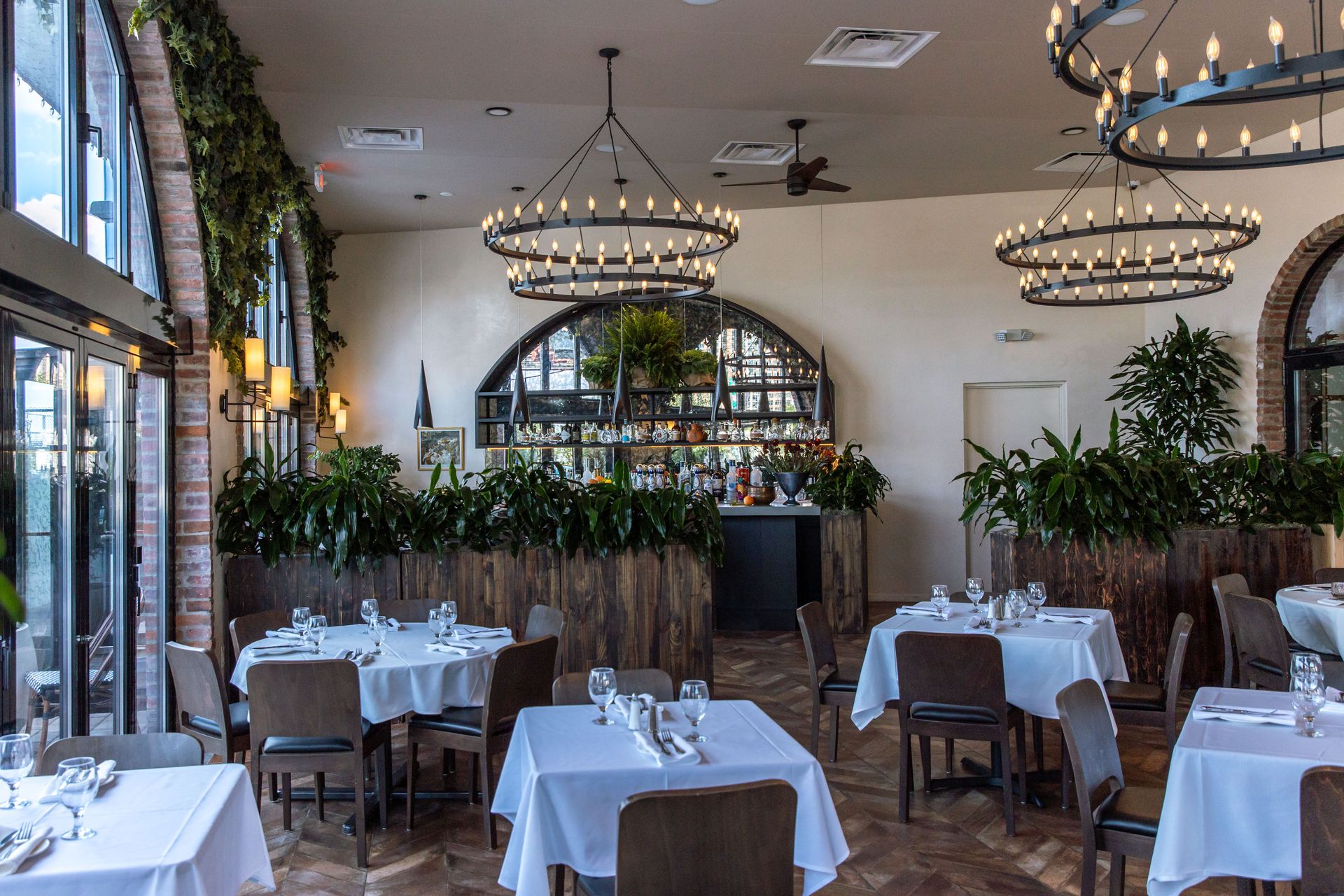 Restaurant interior with tables set for dining, chandeliers, and large windows.