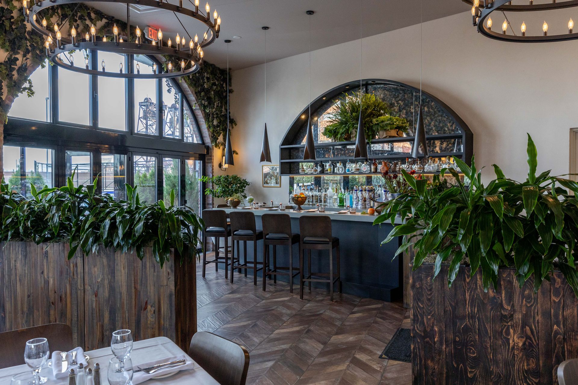 Bar area in a restaurant with large windows, plants, and high-top seating. Dark wood and ornate lighting.