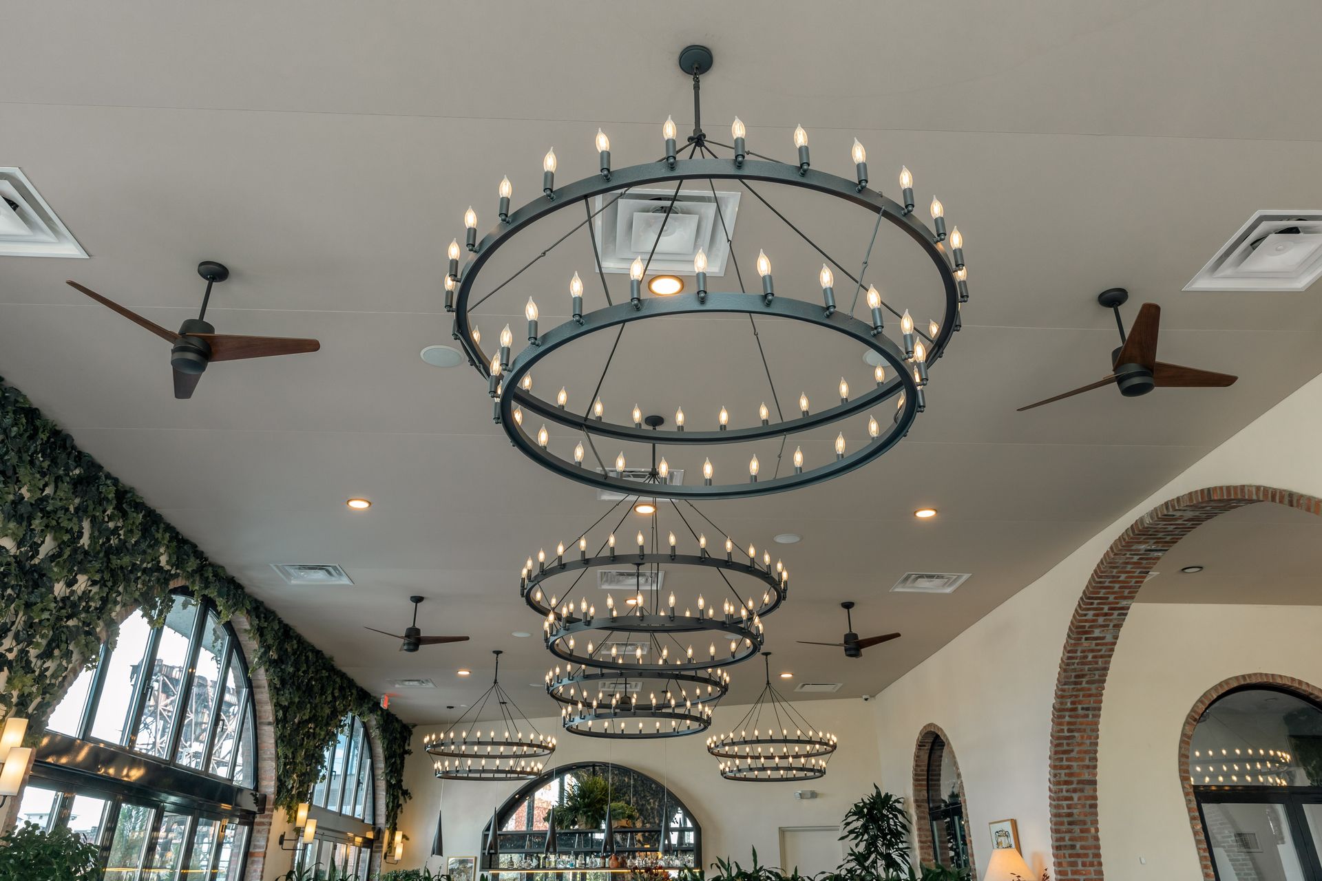 A restaurant interior with large circular chandeliers and ceiling fans. Arched windows with greenery.