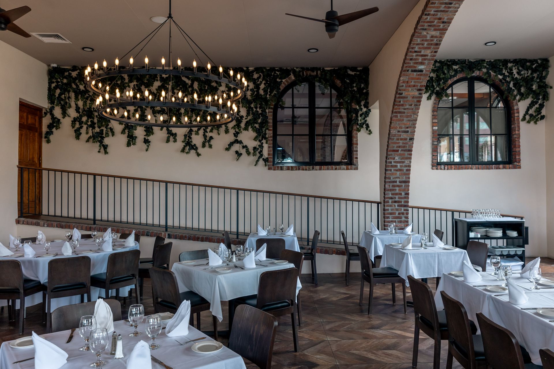 Restaurant dining area with tables set for a meal, a large chandelier, and arched windows with ivy.