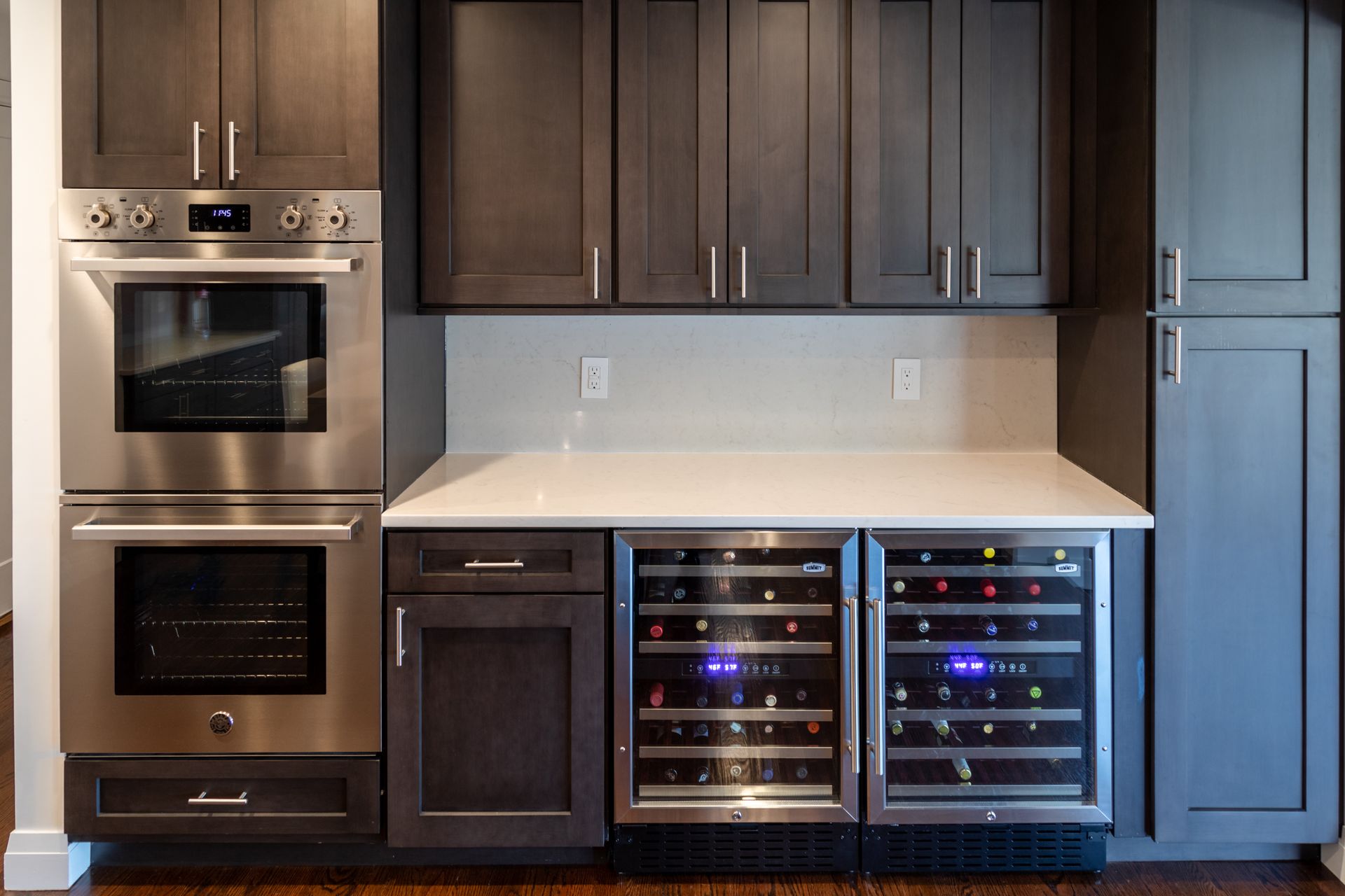 Modern kitchen cabinets with built-in oven, wine coolers, and countertops. Dark wood cabinets with stainless steel appliances.