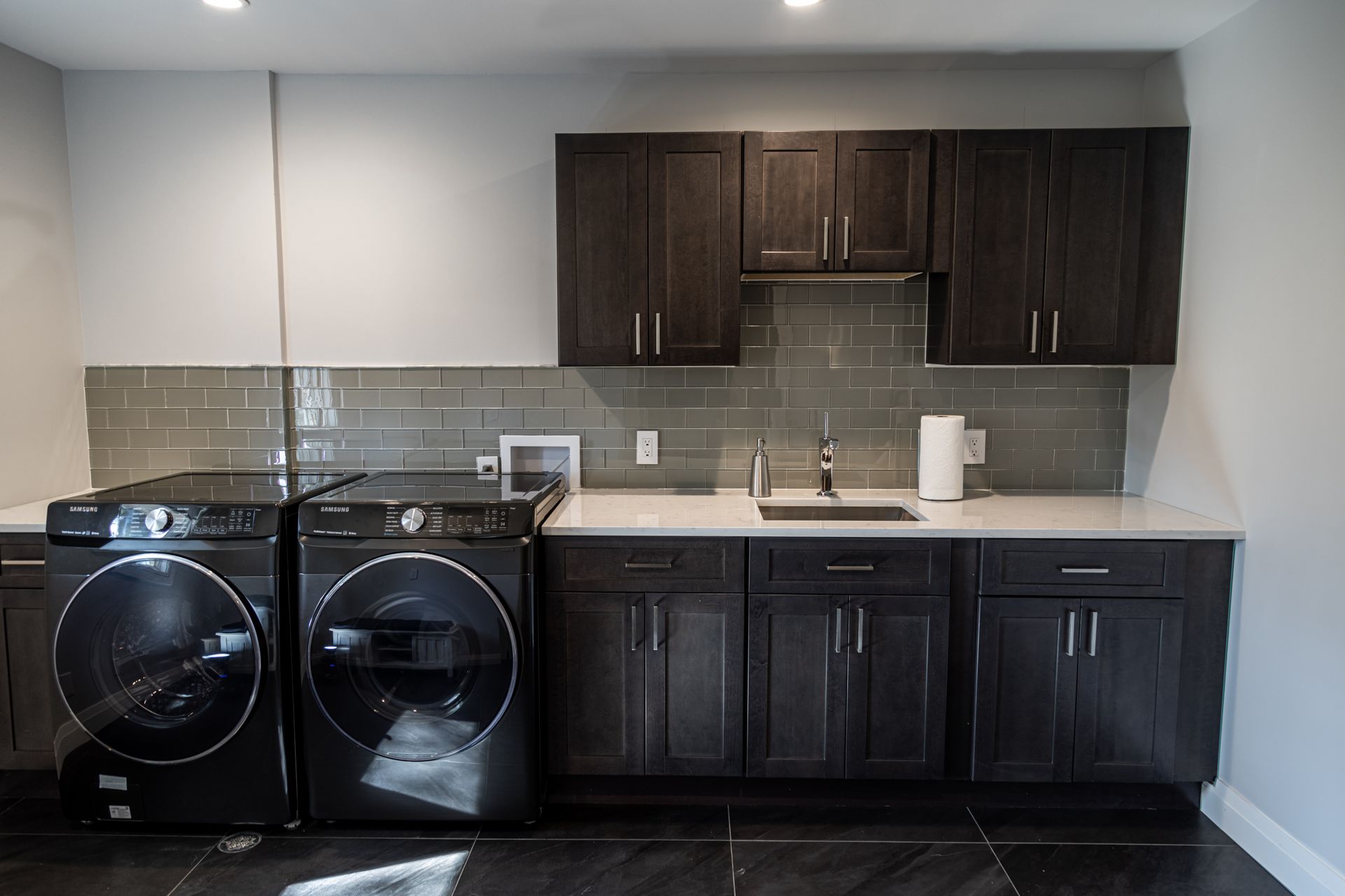 Laundry room with dark cabinets, gray backsplash, and black washer and dryer.