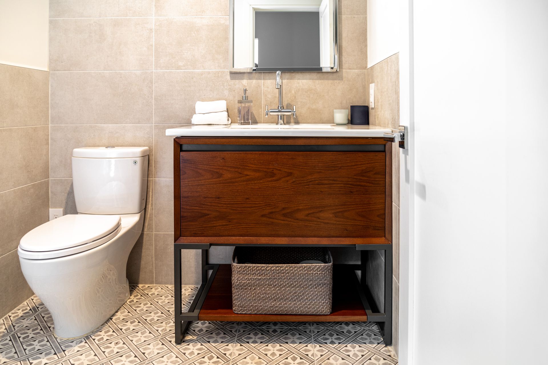 Bathroom with toilet, vanity, and basket under the sink. Beige tile walls and stone floor.