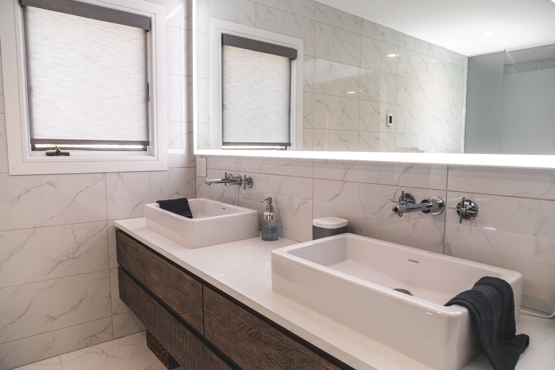 Bathroom with two white sinks on a wooden vanity, marble walls, and a large mirror.