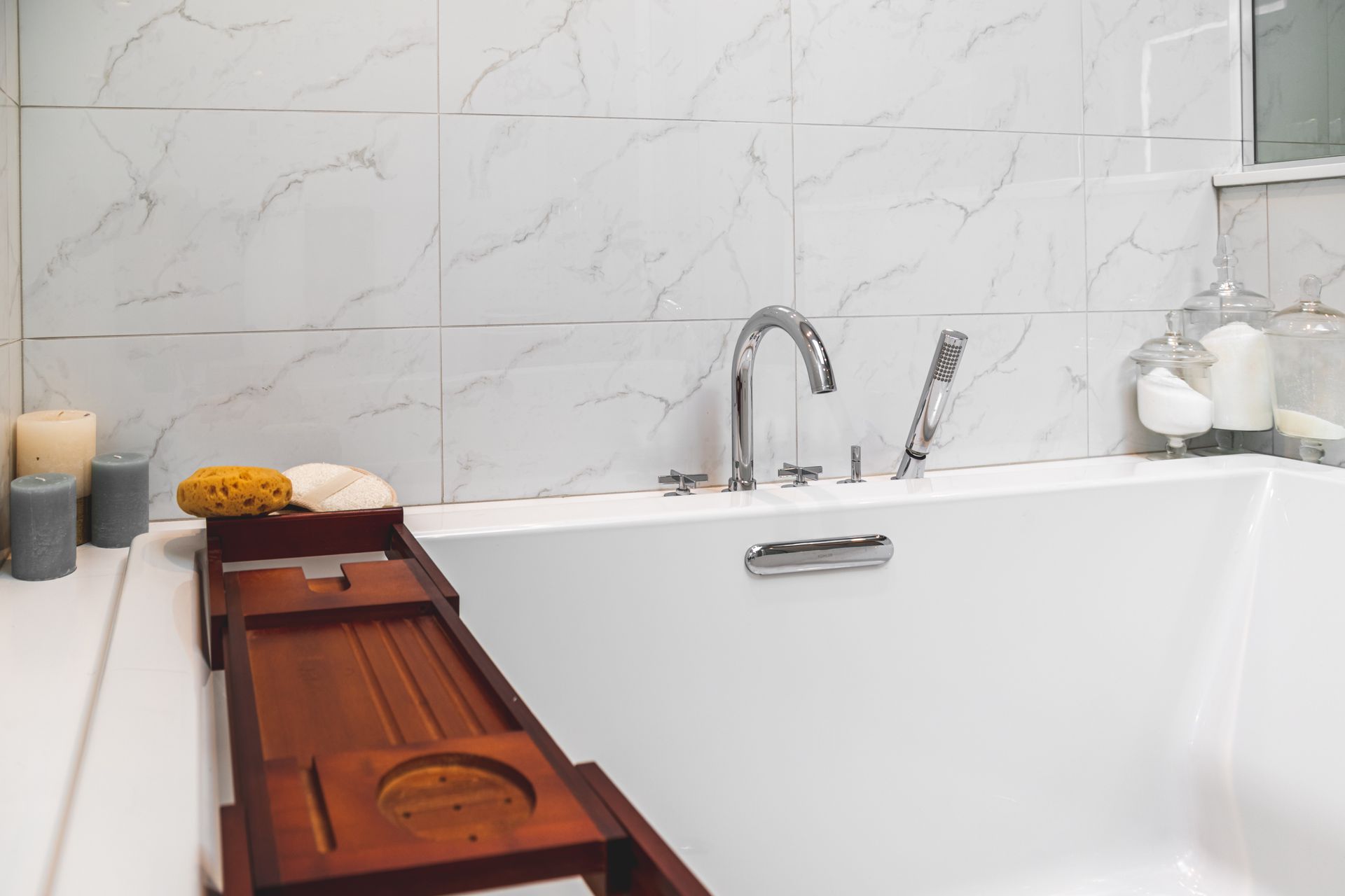 White bathtub with chrome fixtures; marble tile background. A wooden bath tray holds a sponge.