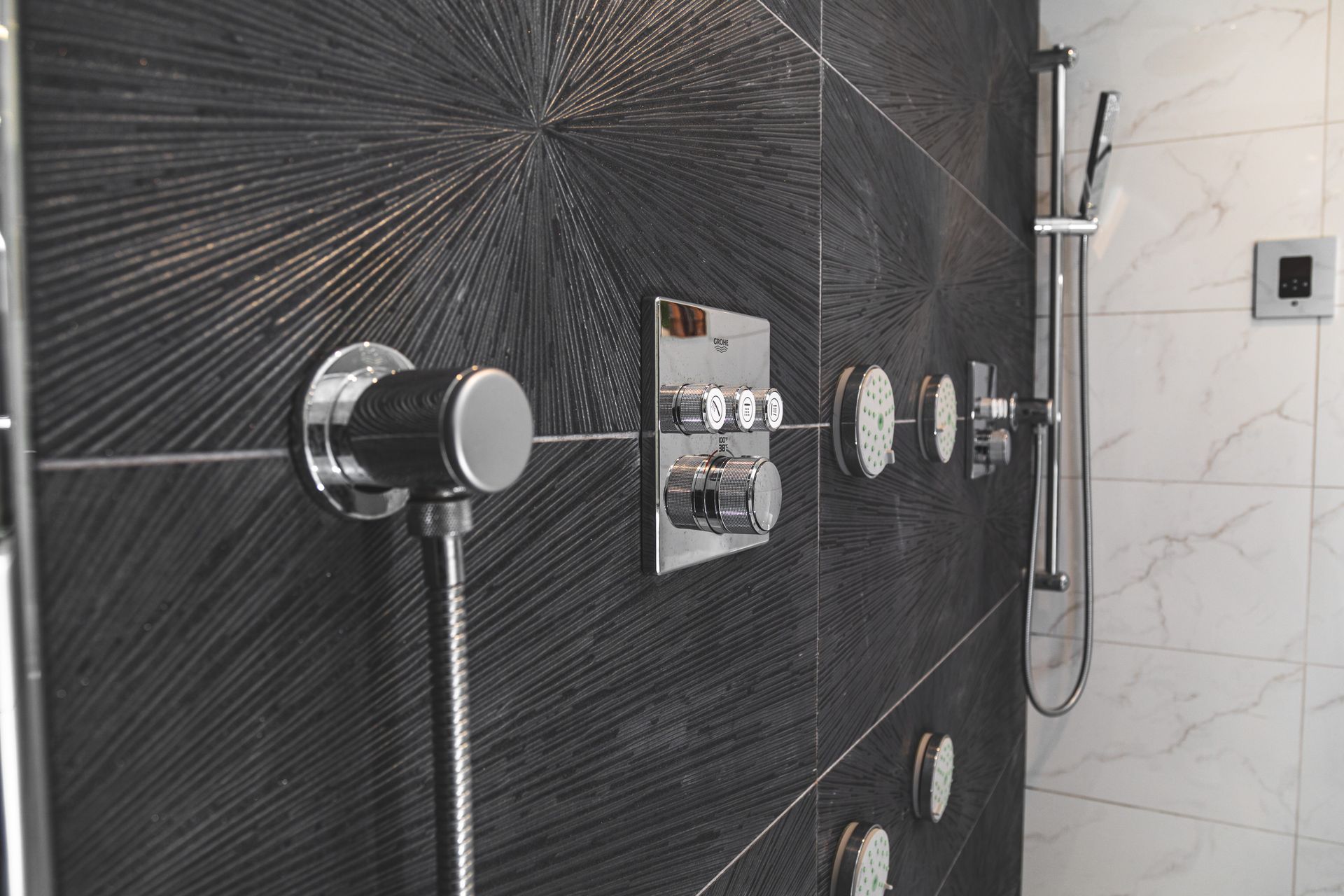 Black and white tiled shower wall with chrome fixtures. Radial pattern on black tile, hand shower, controls.