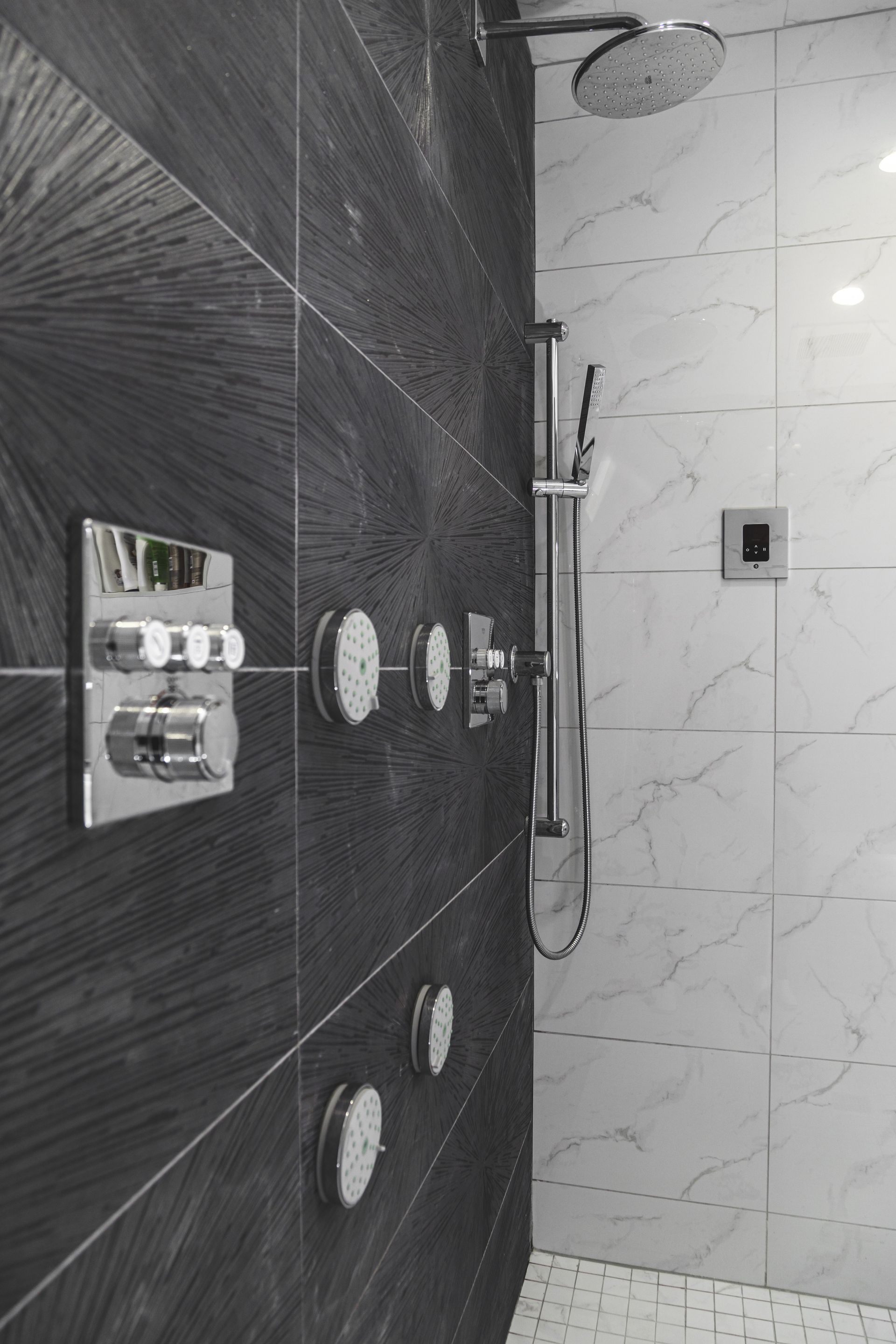 Black and white shower with multiple showerheads and controls on dark and light tiled walls.