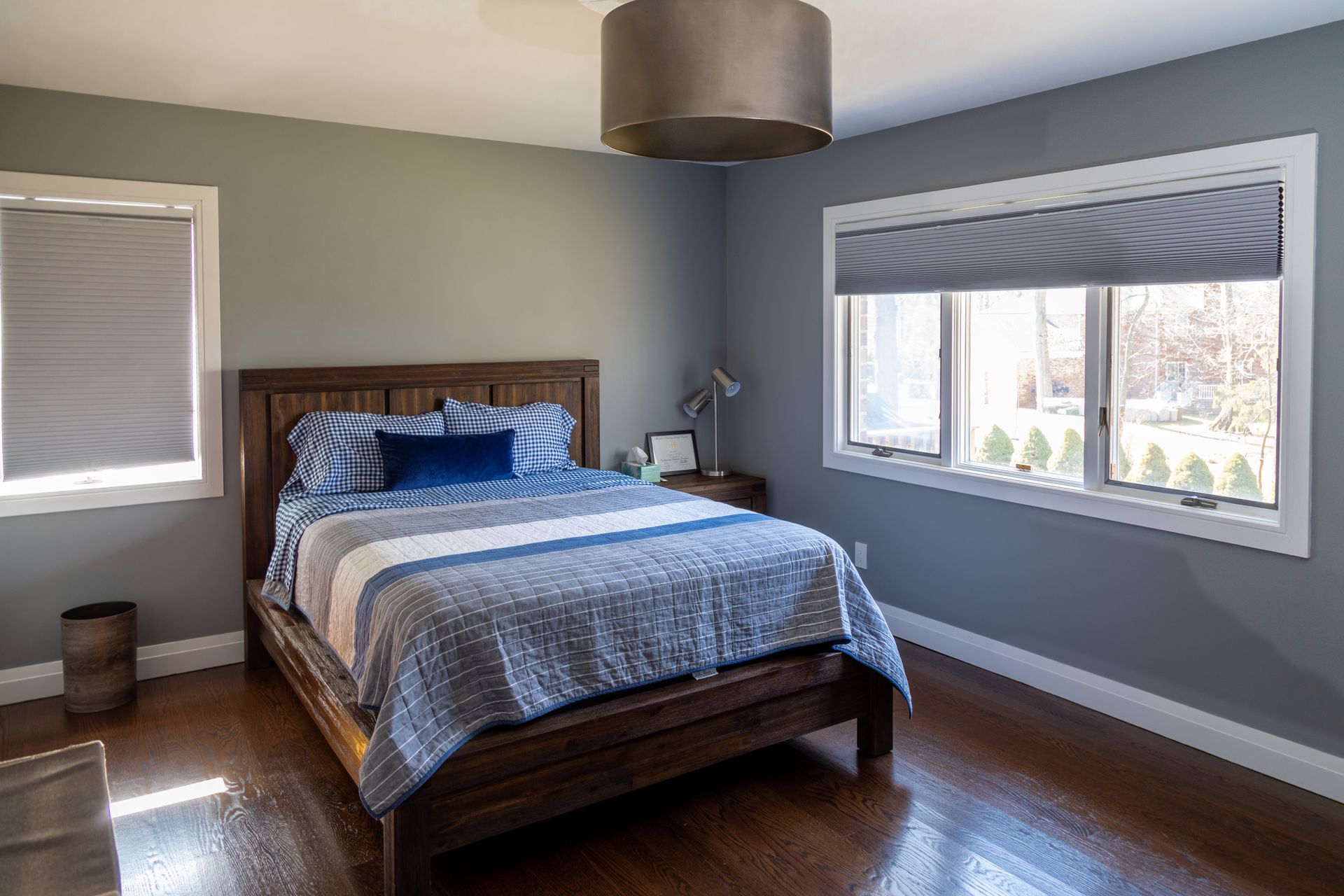 Bedroom with a wooden bed, blue and white bedding, two windows with blinds, and a gray wall.