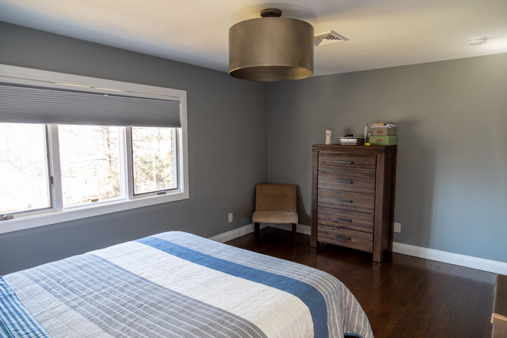 Bedroom with gray walls, bed with blue and white striped quilt, wooden dresser, small chair, and a window.