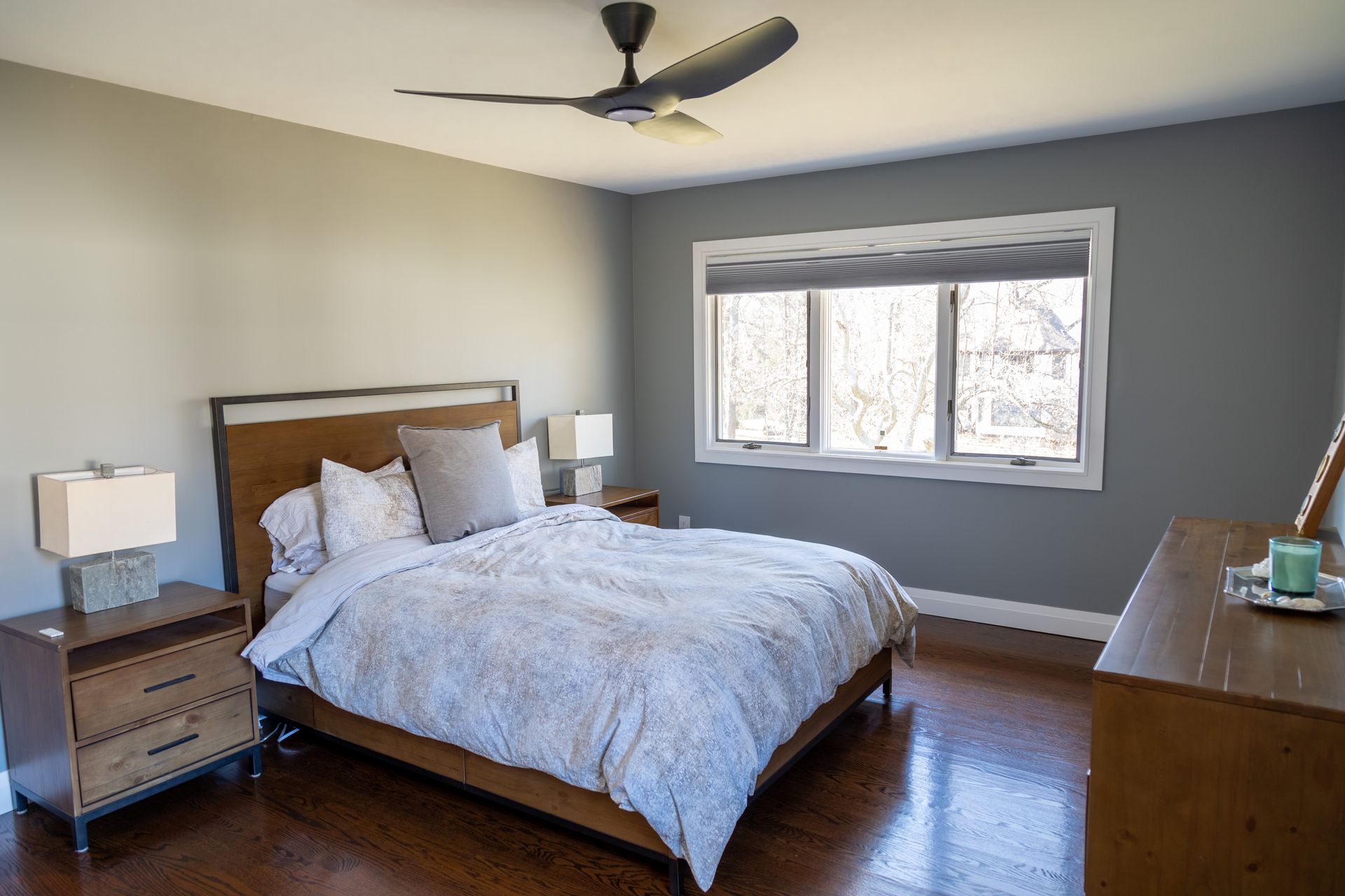Bedroom with bed, nightstands, dresser, and window. Gray walls, brown floors, and a ceiling fan.