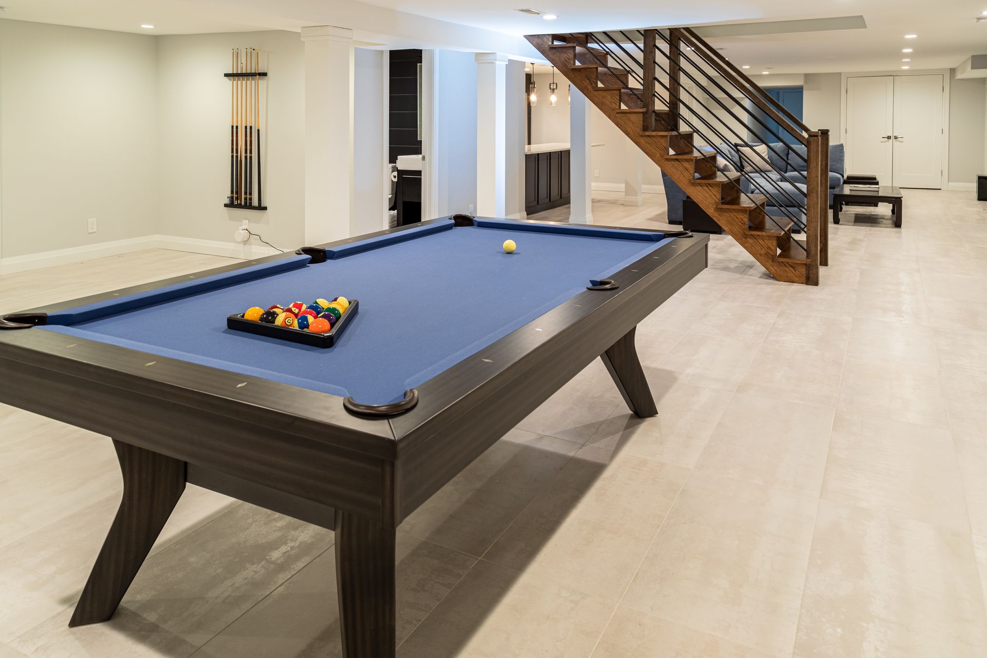 Pool table in a finished basement with stairs, blue felt, colorful balls, and hardwood floors.