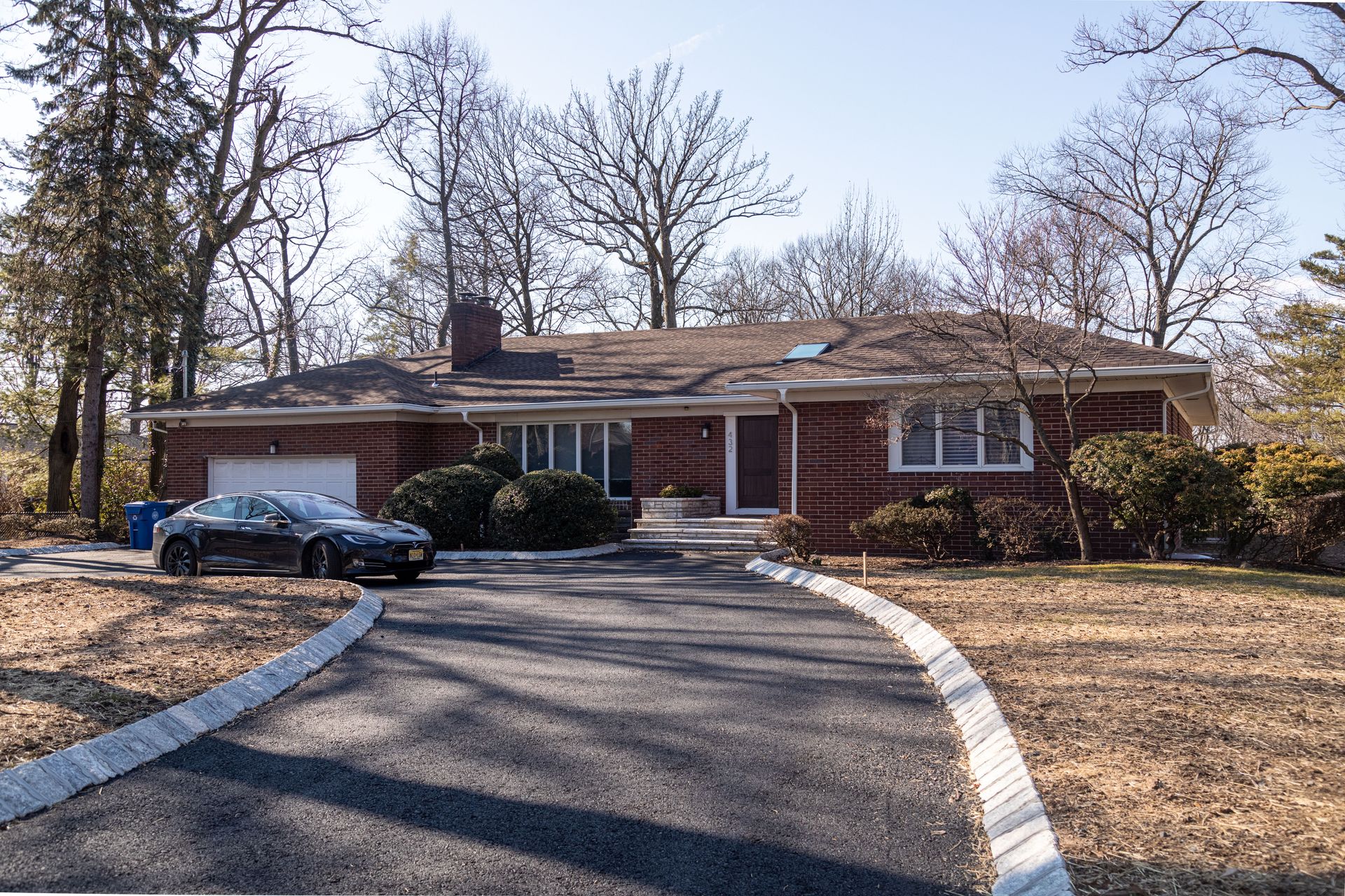 House with a long driveway; a black car is parked in front of a garage. Trees surround the house.