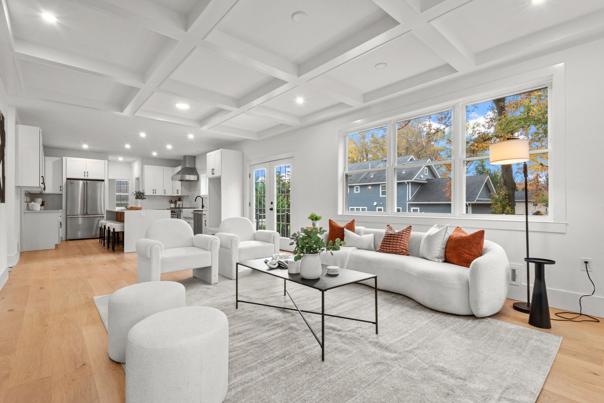 Bright living room with white furniture, wooden floors, and a large window overlooking trees.