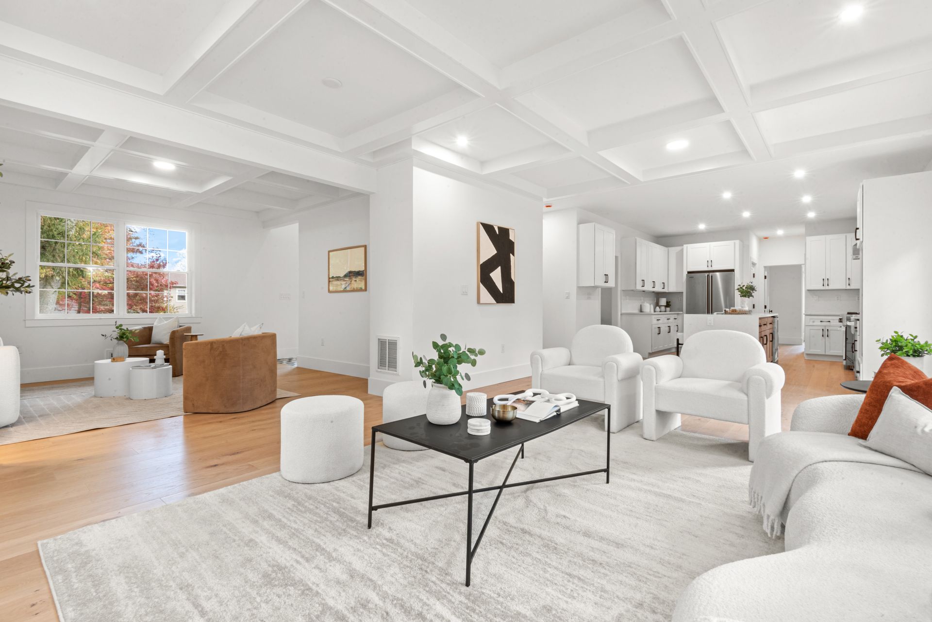 Bright, open-concept living room with white walls, wood floors, and neutral furniture; kitchen in the background.