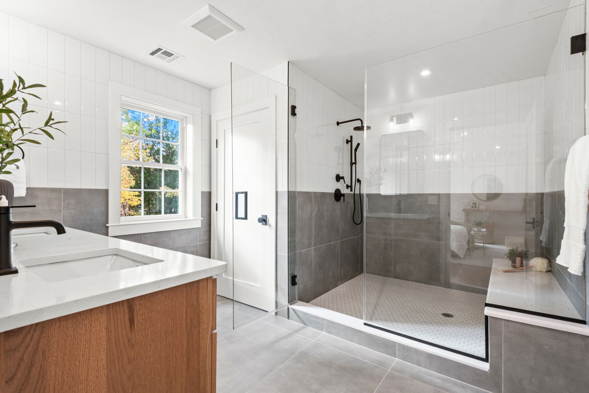 Modern bathroom with a glass shower, white and gray tile, wooden vanity, and a window.