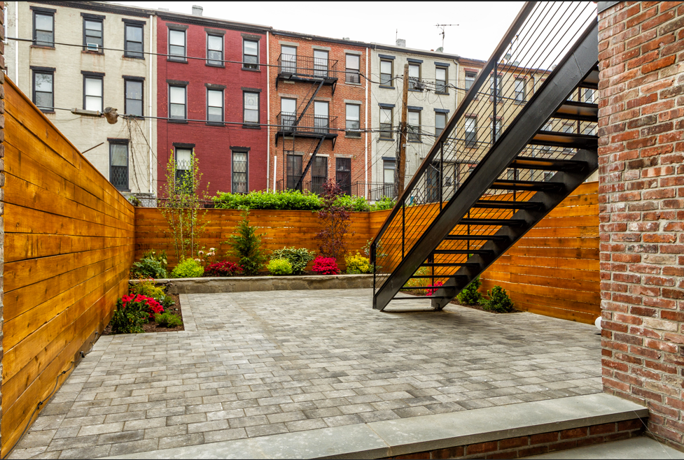 Backyard patio with wooden fence, brick building, and metal staircase leading to colorful townhouses.