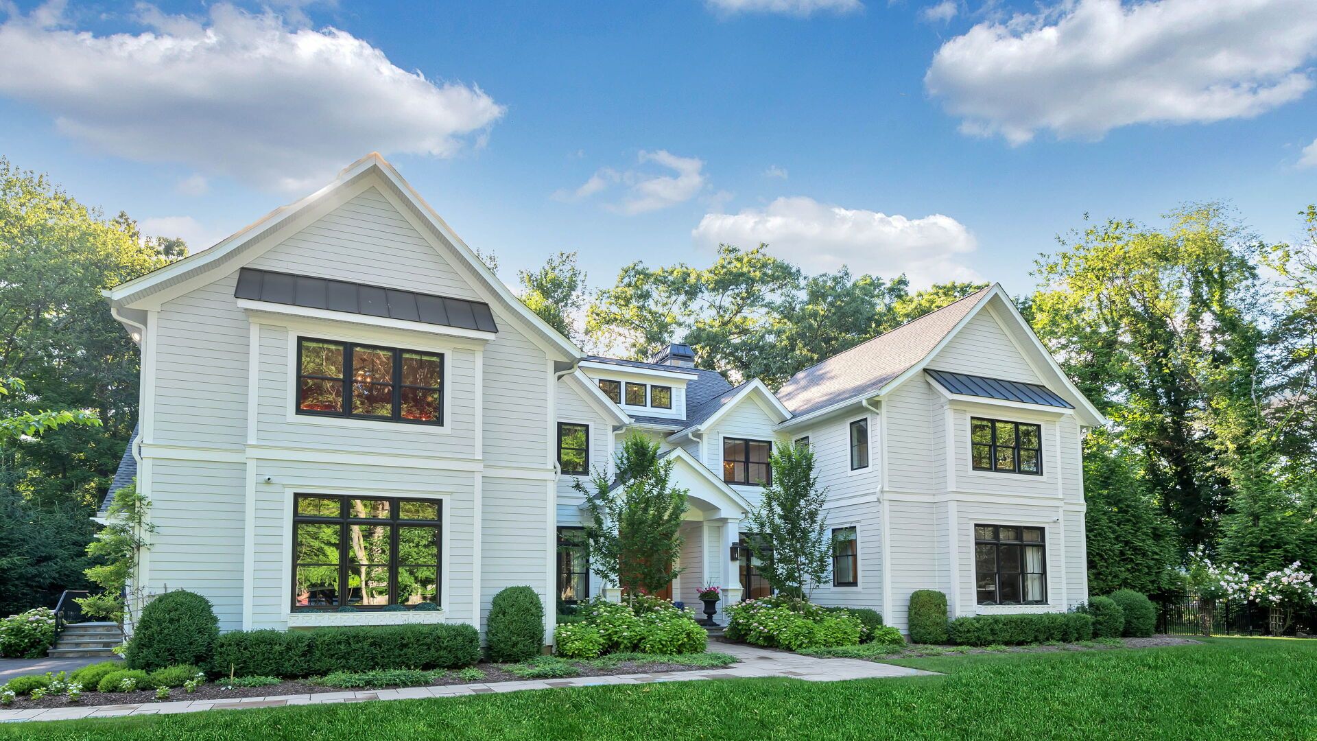 White two-story house with black-framed windows, surrounded by green lawn and trees, blue sky.