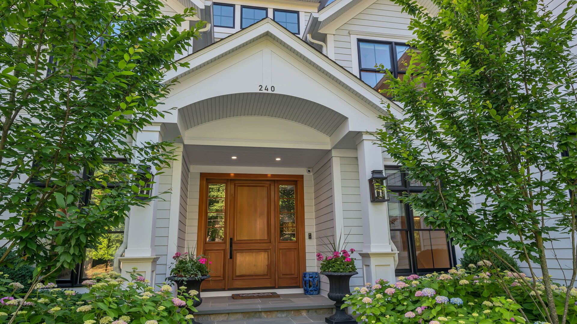 Front entrance of a white house with wooden double doors, flanked by trees and flower pots.