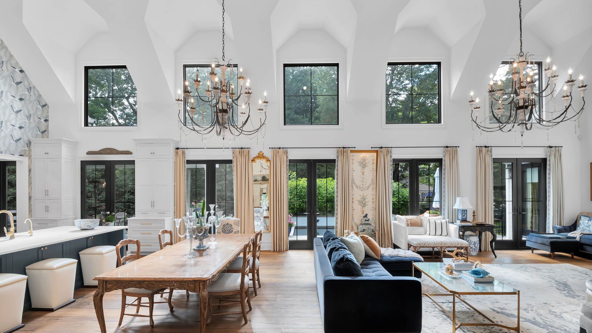 Elegant living room with high ceilings, chandeliers, large windows, and a long dining table.