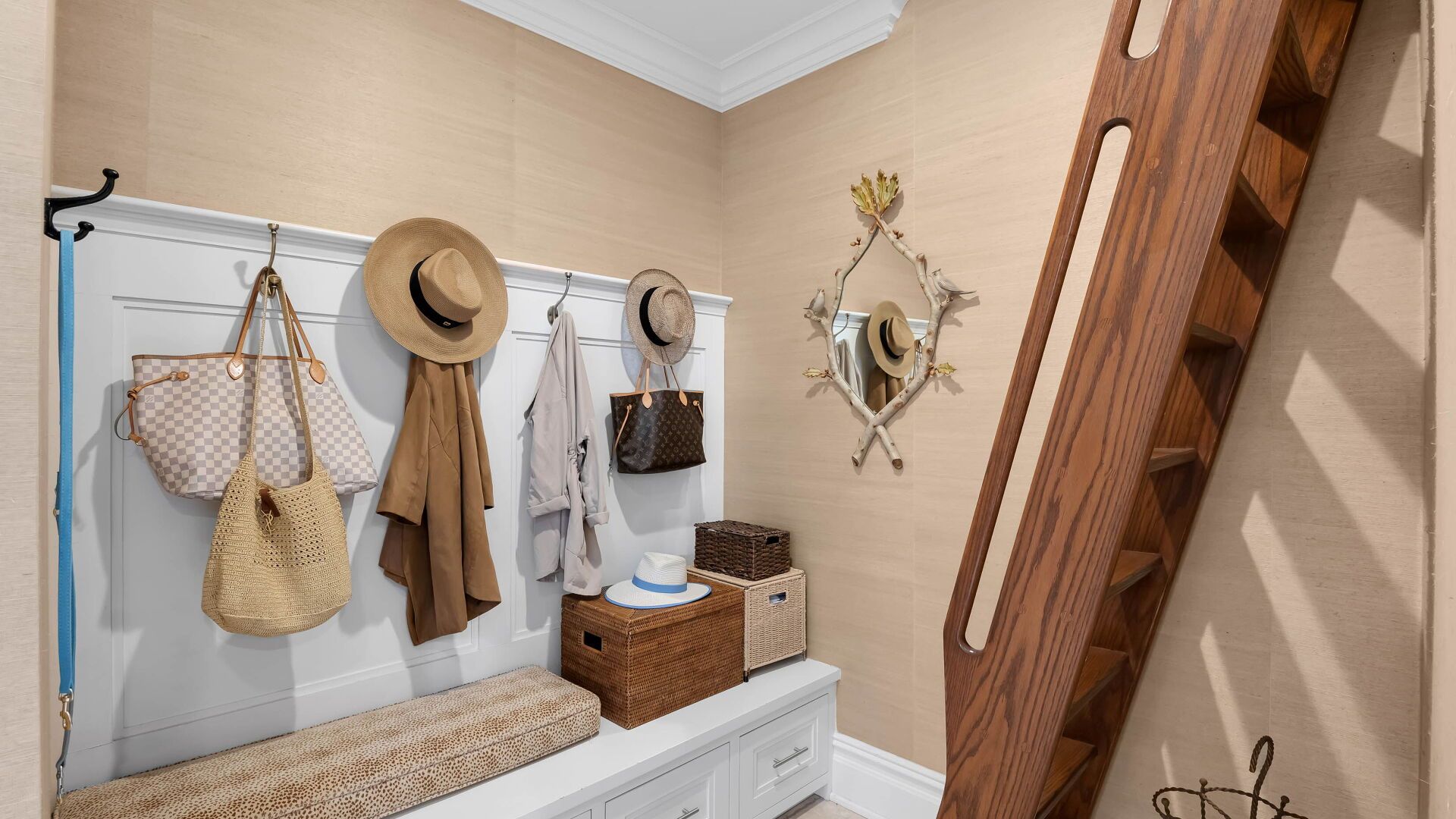 Entryway with white bench, hooks with hats and bags, brown woven storage, and a wooden staircase.