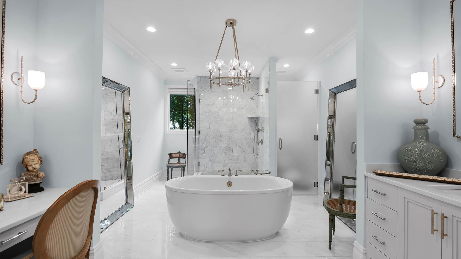 Elegant, light-blue bathroom with a freestanding tub, chandelier, marble shower, vanity, and large mirrors.