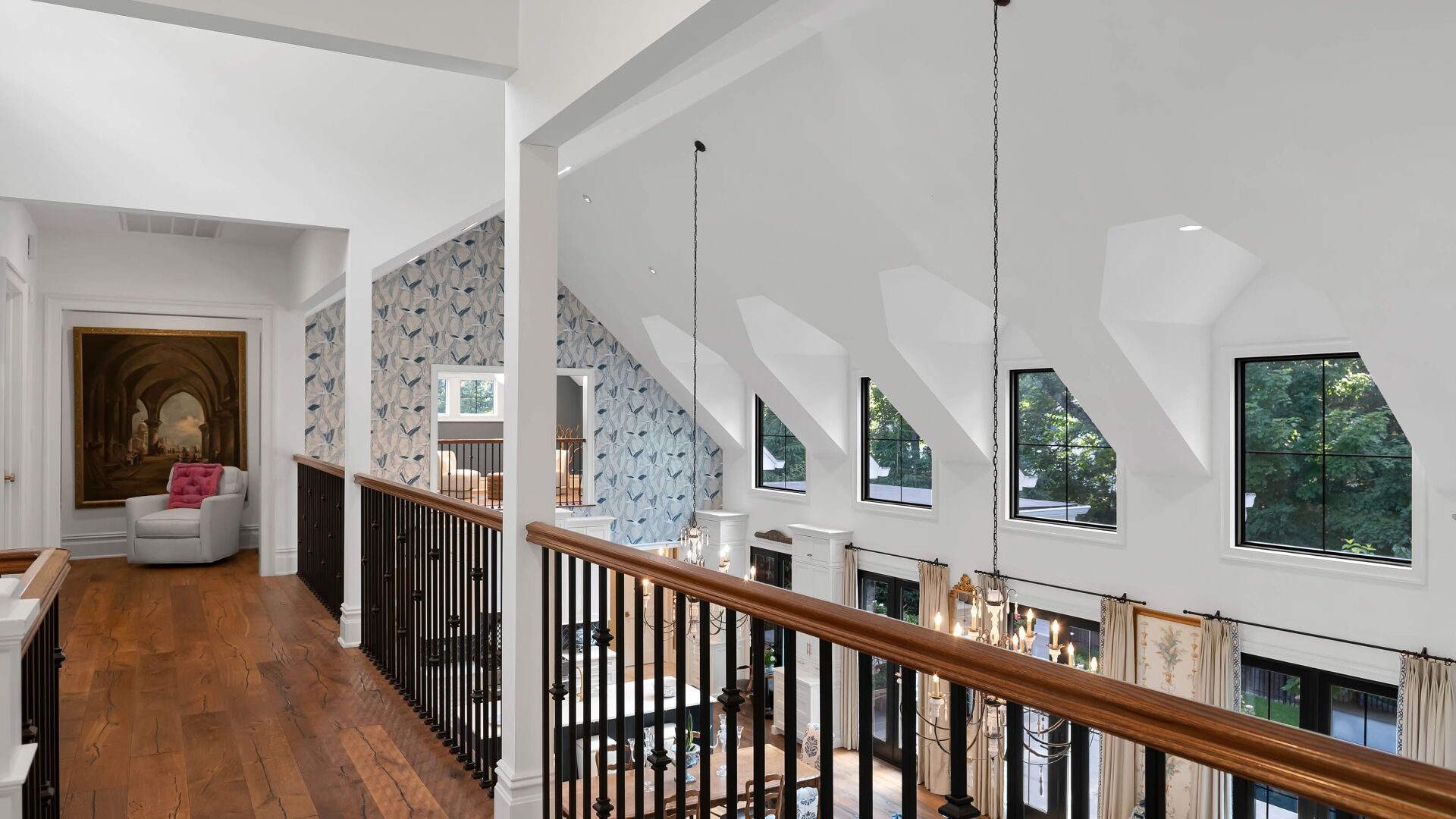 Wooden hallway with high ceilings, windows, and black railings overlooking a chandelier.