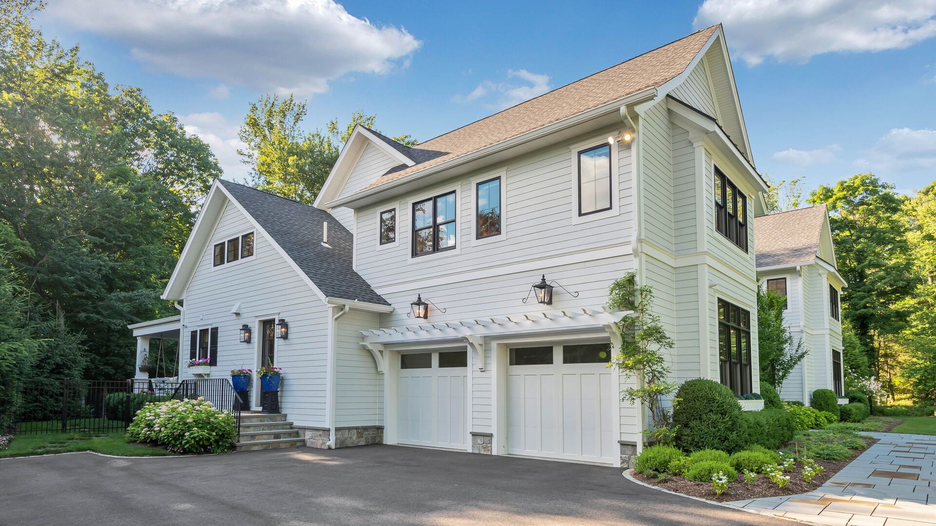 White two-story house with two-car garage, dark trim, and paved driveway. Sunny day with trees in the background.