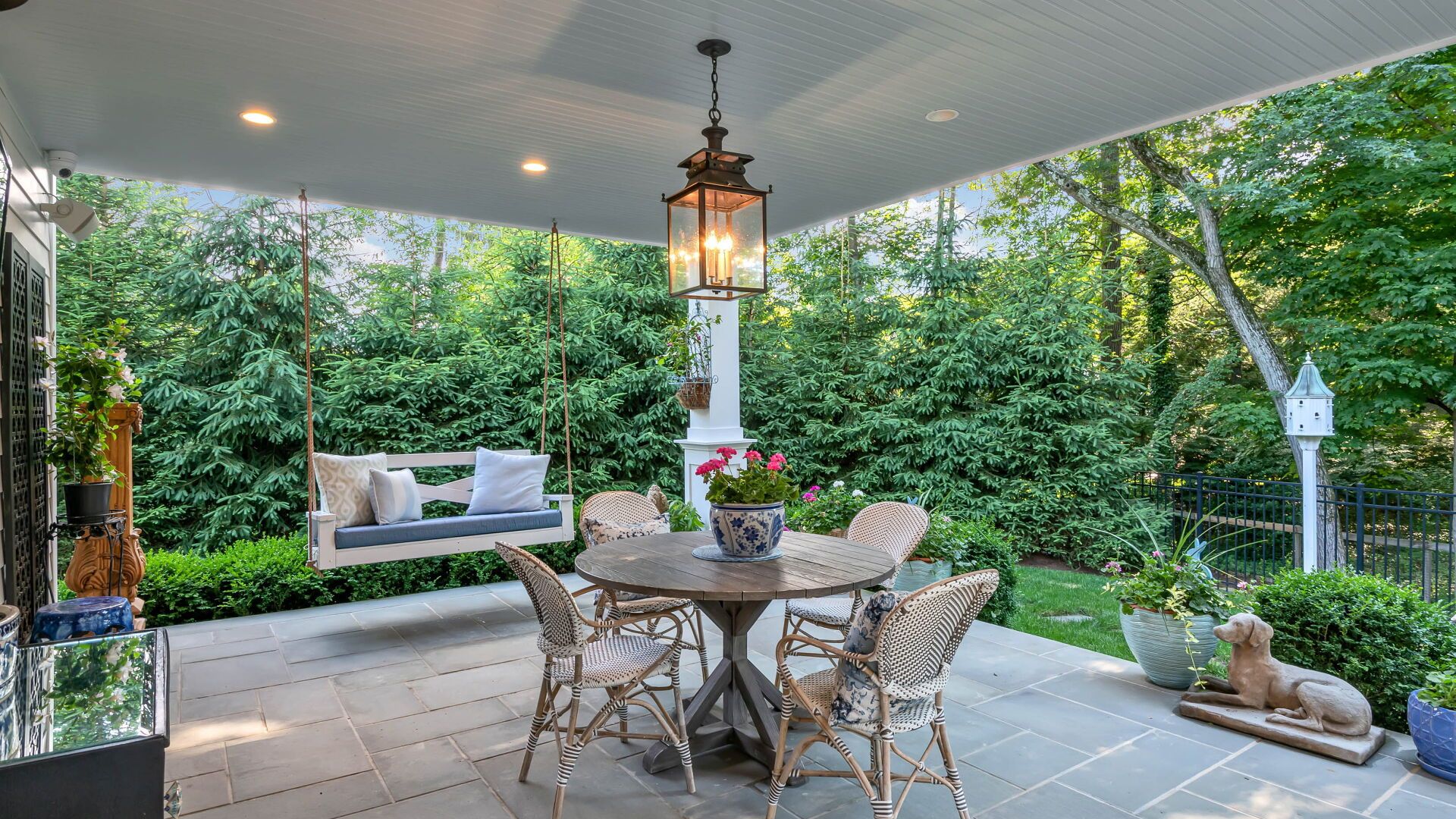 Covered outdoor patio with a swing, table, chairs, and surrounding greenery.