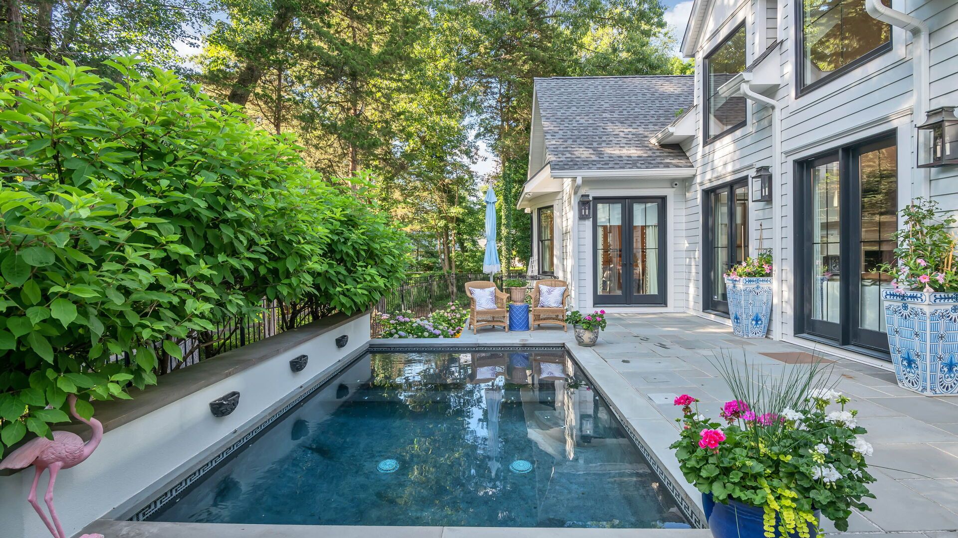 Backyard pool and patio area. White house, greenery, blue pool, chairs, and potted flowers.
