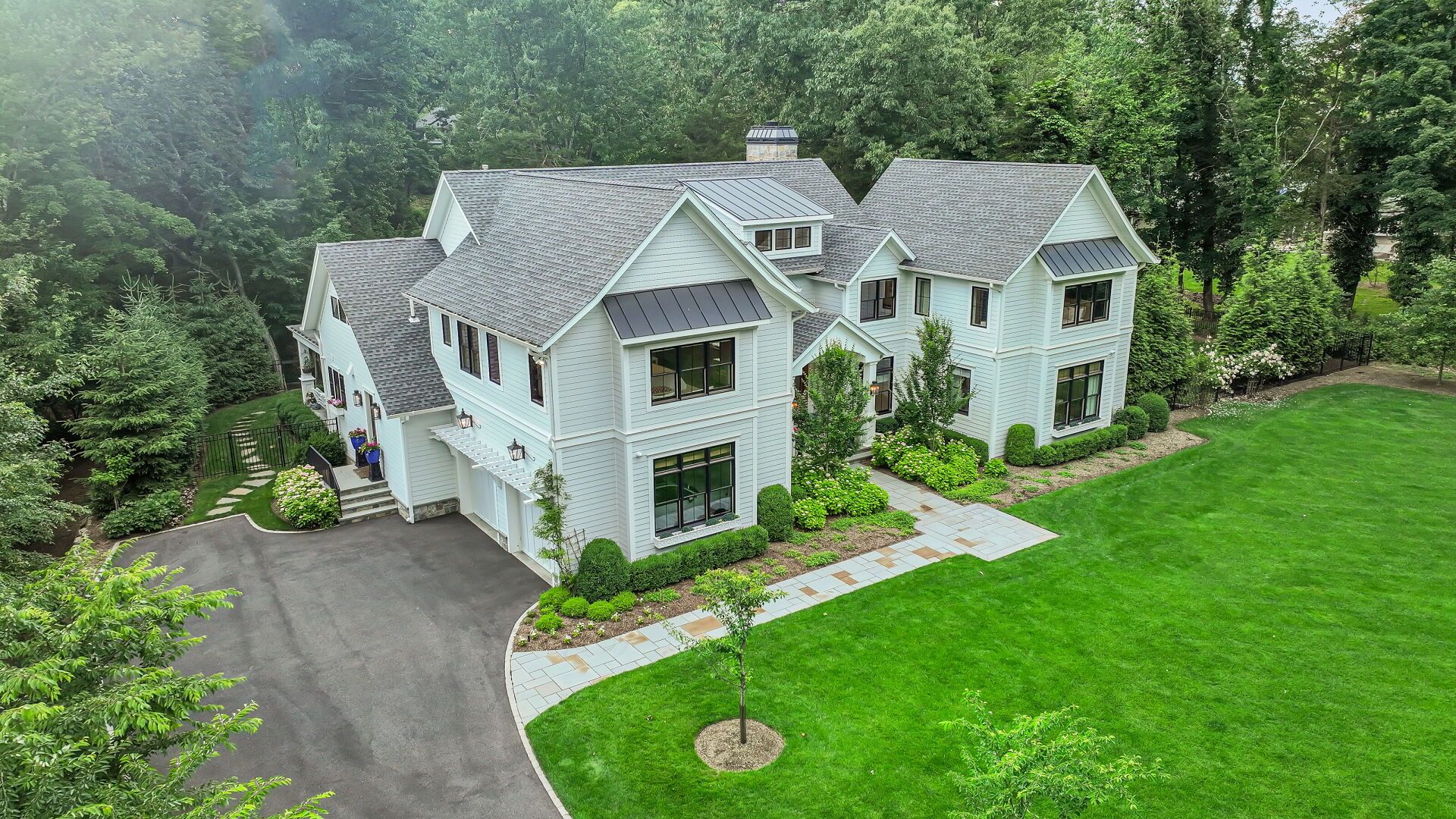 White farmhouse with gray roof, surrounded by green lawn and trees. Driveway in foreground.