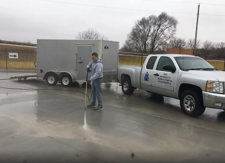 A man is cleaning a concrete driveway next to a truck and trailer.