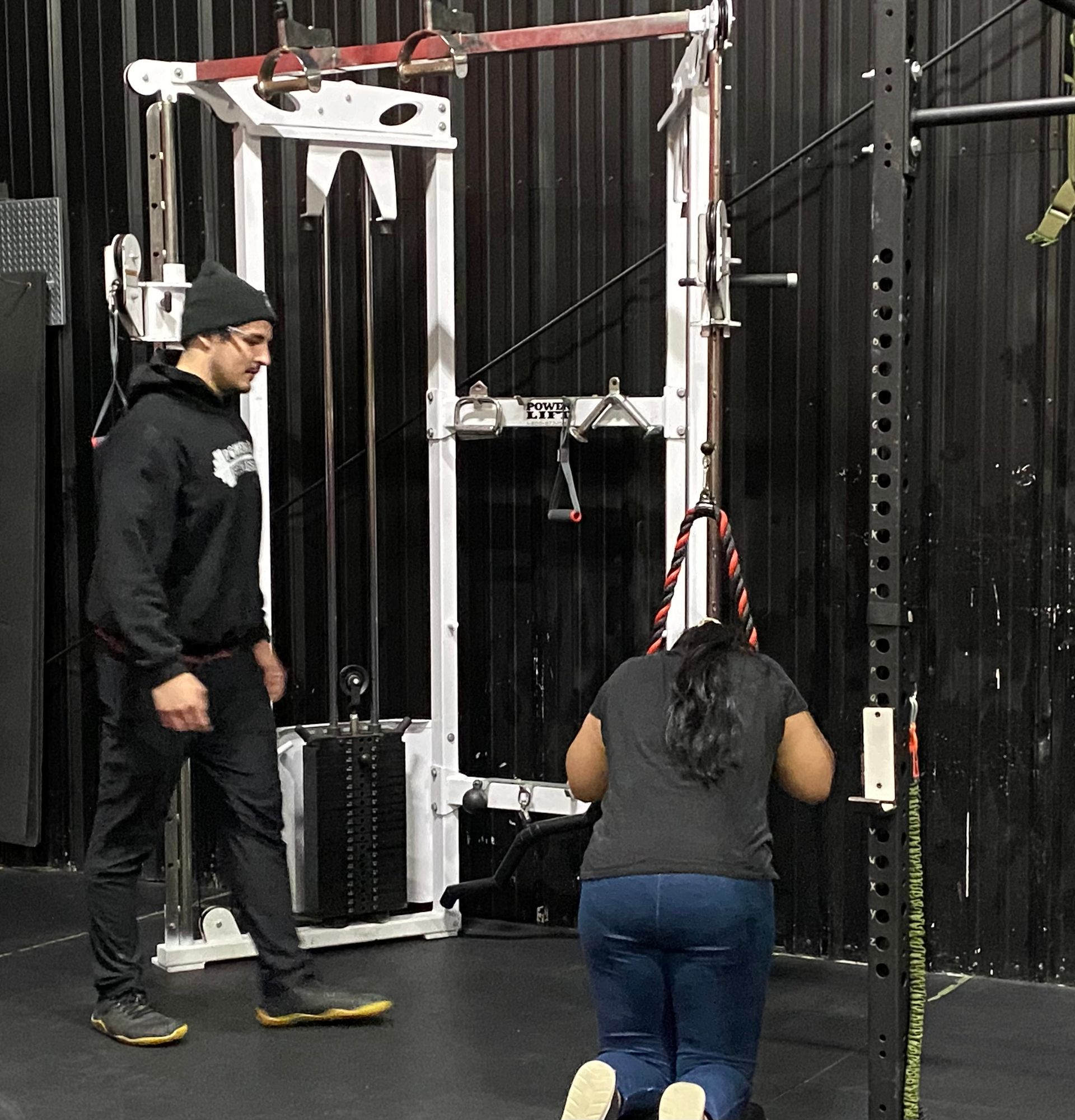 Two people in a gym, one kneeling by a cable machine while another stands nearby.