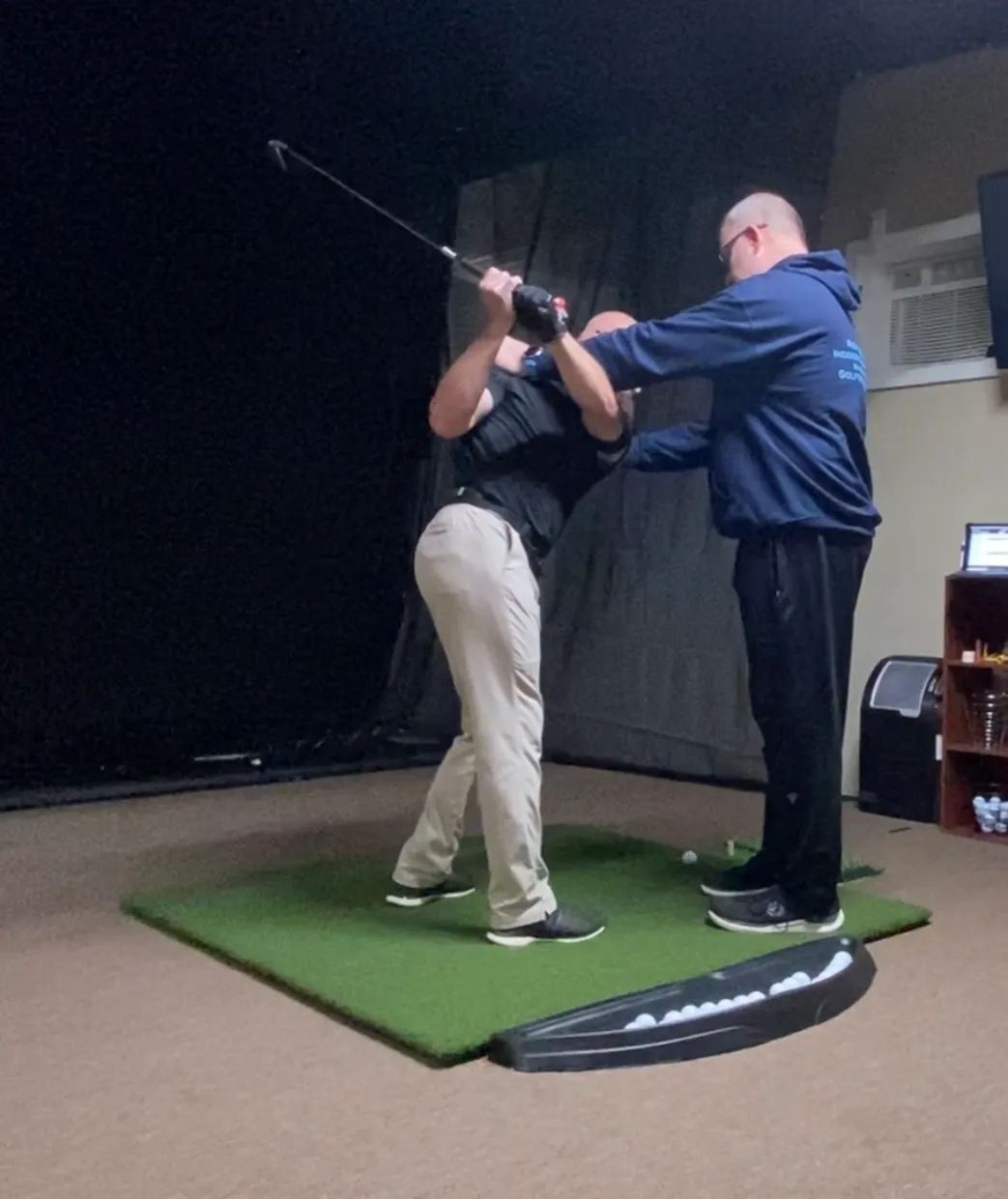 Golf instructor helping a student practice a swing on an indoor mat.