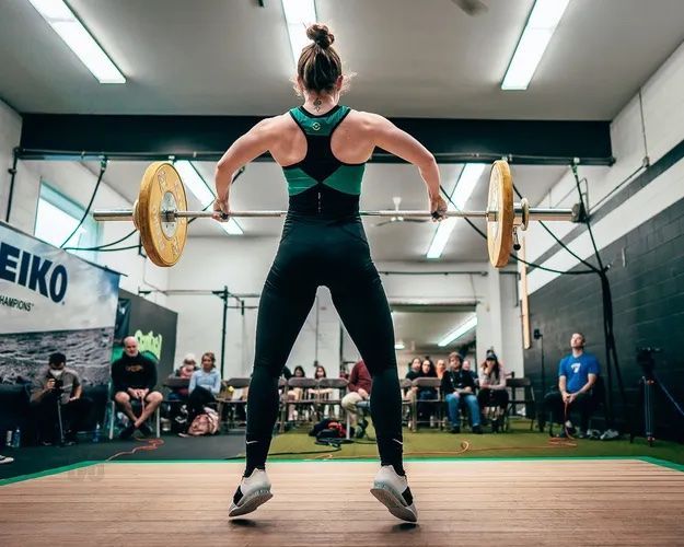 Weightlifter in black and teal uniform lifting a barbell overhead at an indoor competition