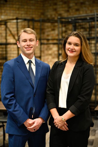 Two people in business attire smiling and standing together indoors against a brick wall.