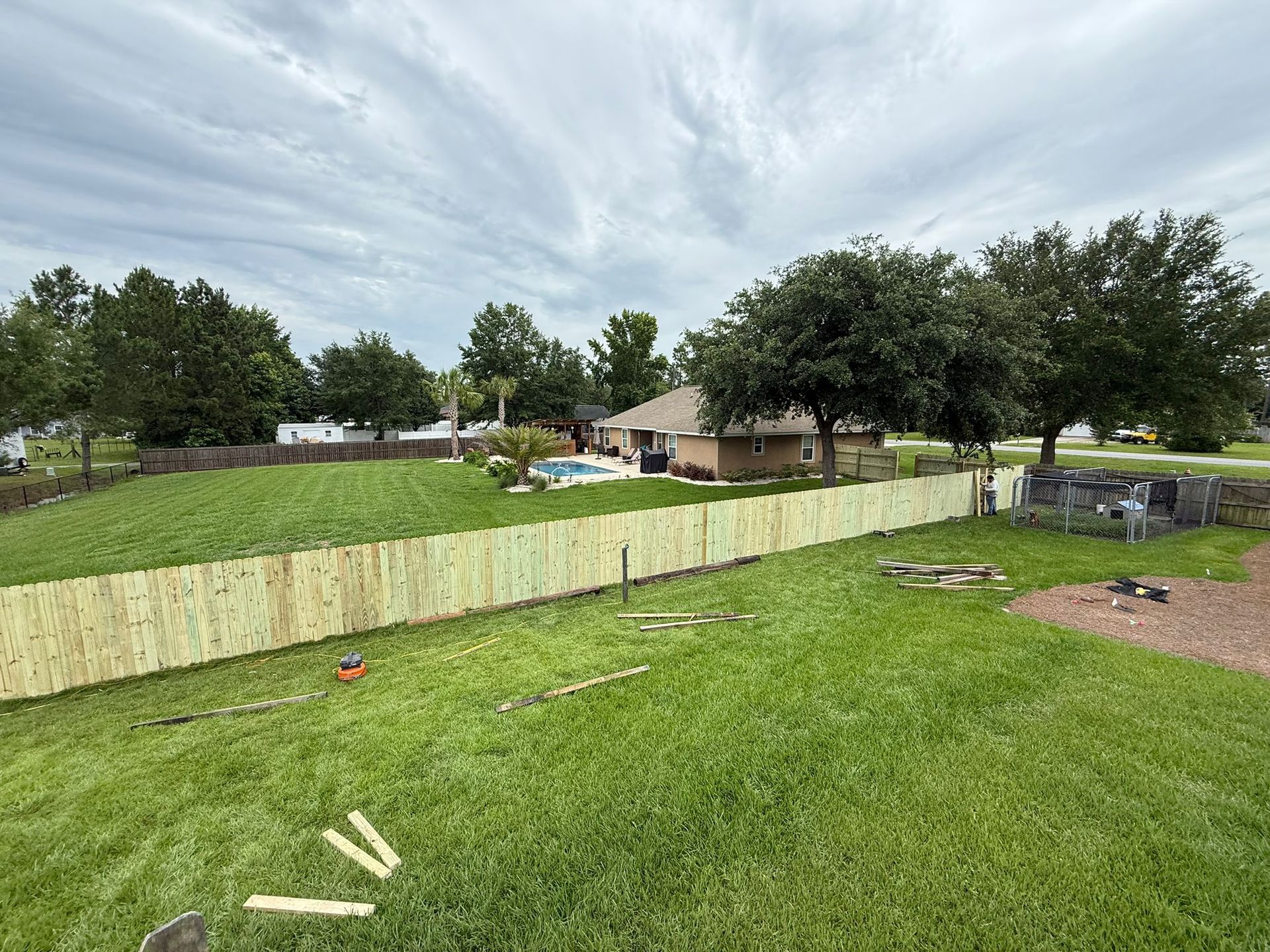 Wooden fence installed in grassy backyard; house with pool in background under cloudy sky.