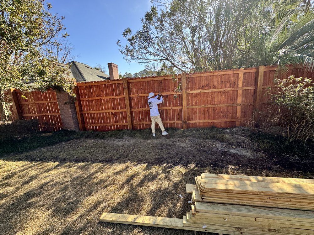 Person staining a new brown wooden fence in a sunny backyard with lumber in the foreground.