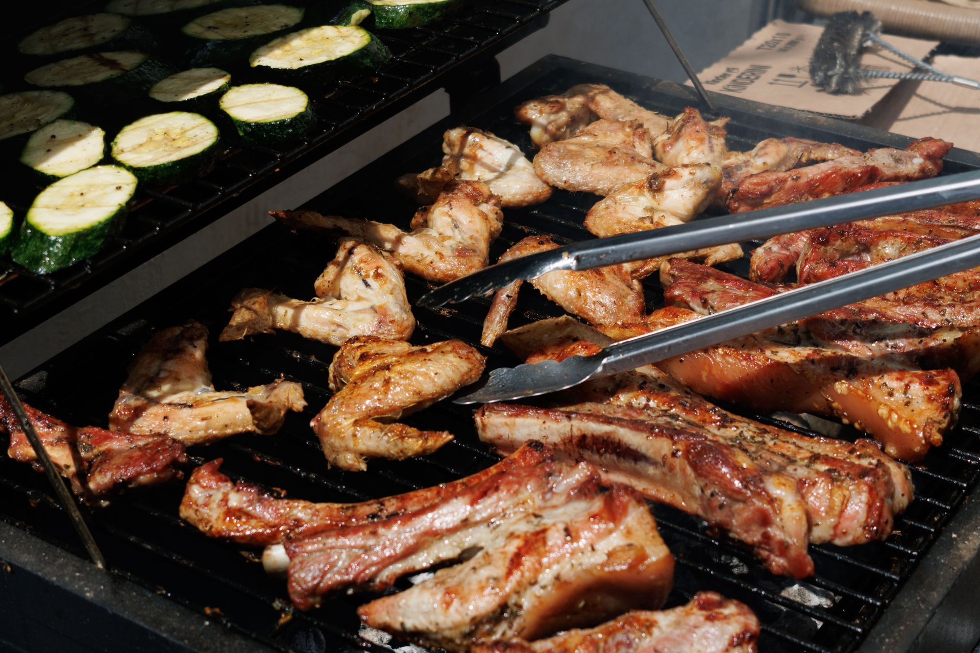 Chicken wings and ribs grilling on a barbecue, with sliced zucchini cooking on the side