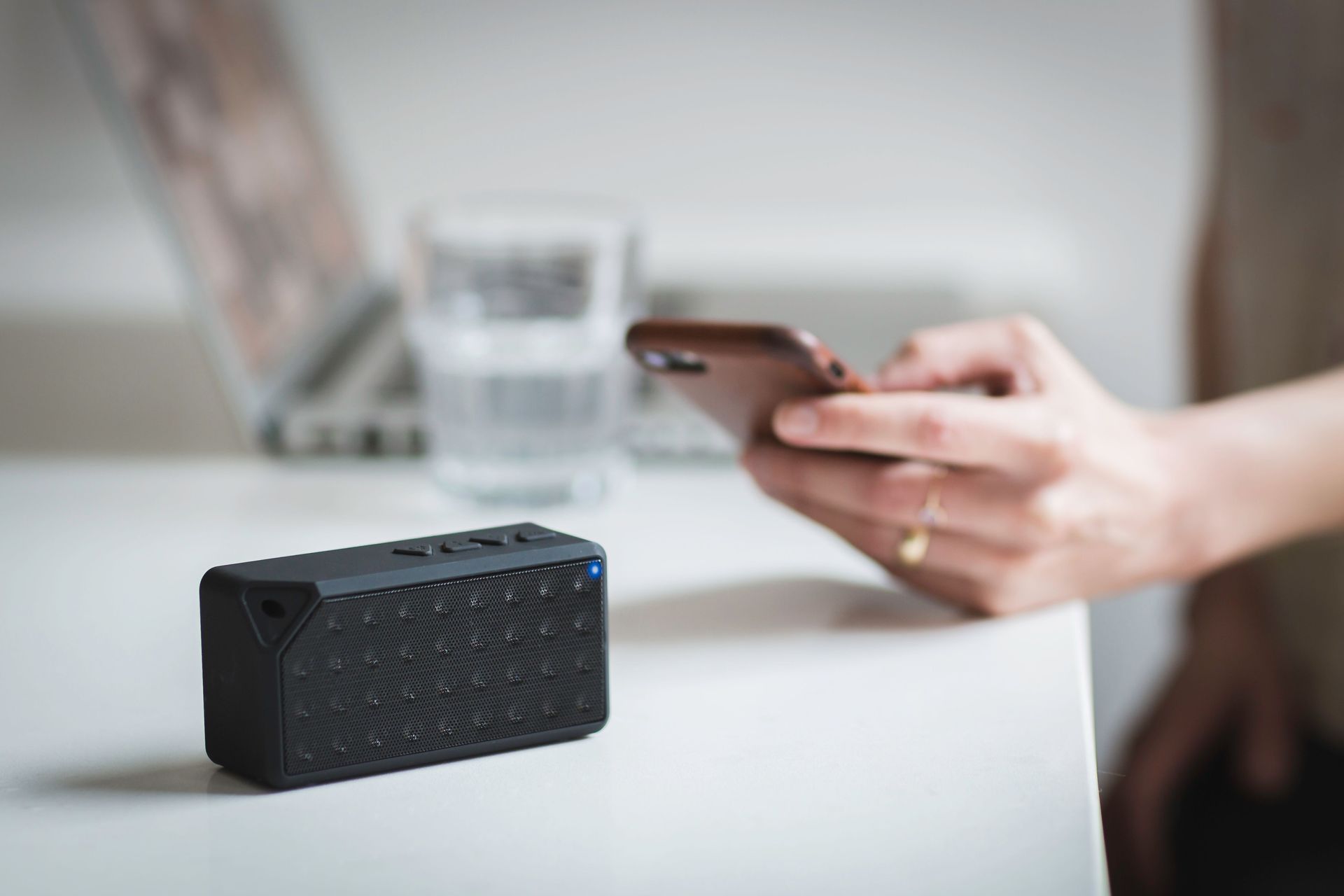 Hand holding a smartphone beside a small black portable speaker on a white table