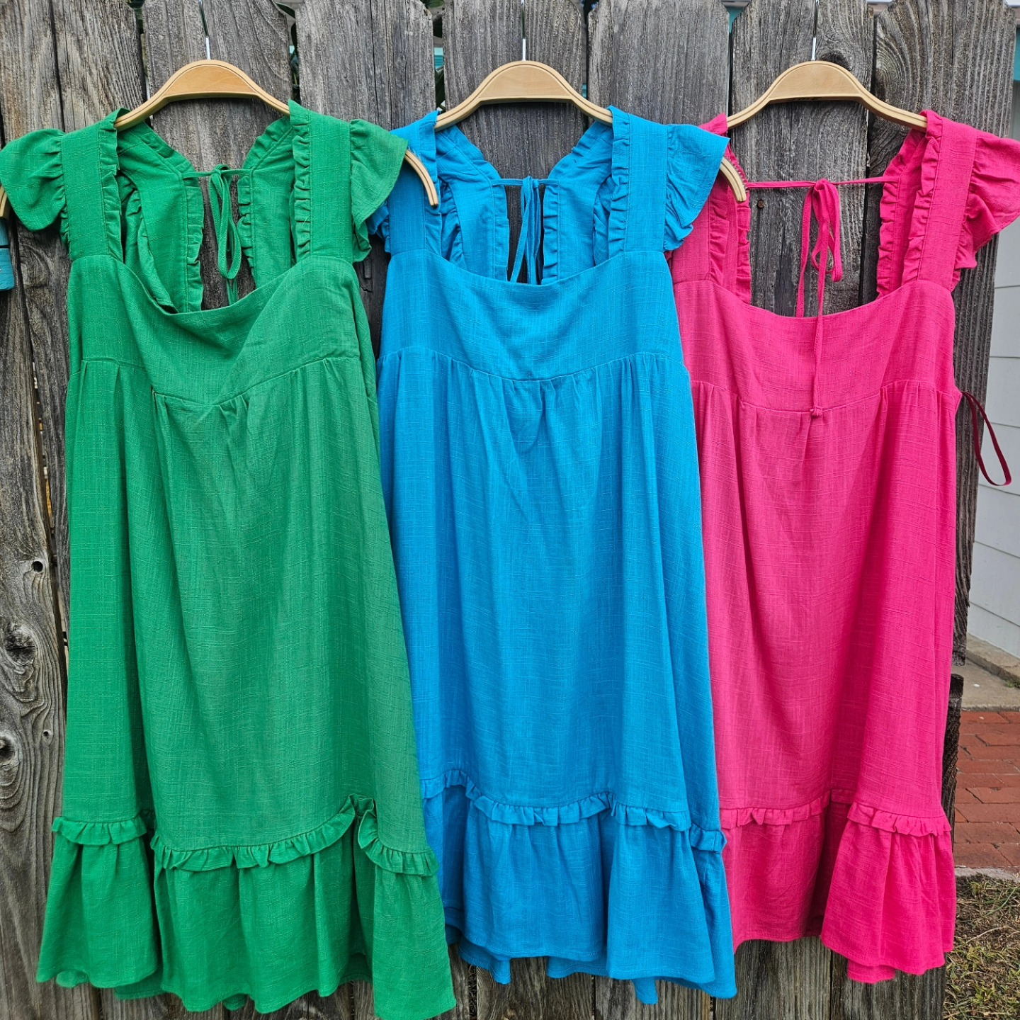 Three colorful sleeveless dresses—green, blue, and pink—hang on wooden hangers against a weathered wood fence.