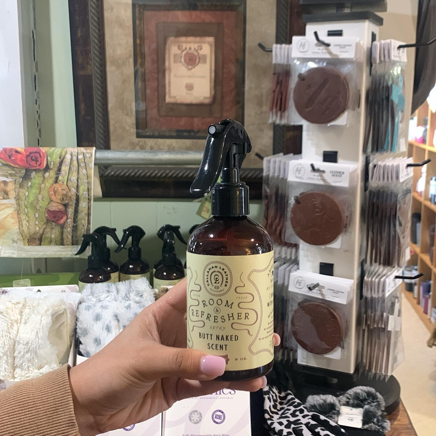 Hand holding a spray bottle in a store aisle with packaged hair accessories and framed decor in the background