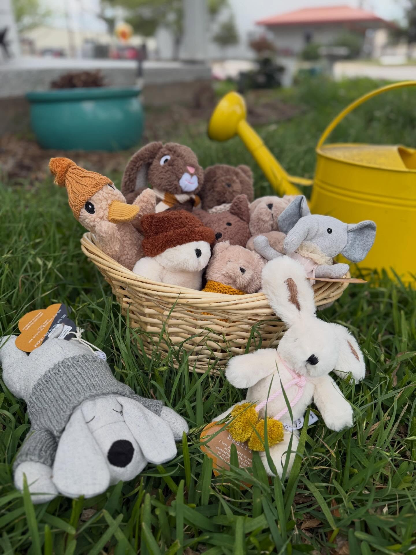 Basket of stuffed animal toys in grass, with a yellow watering can nearby.