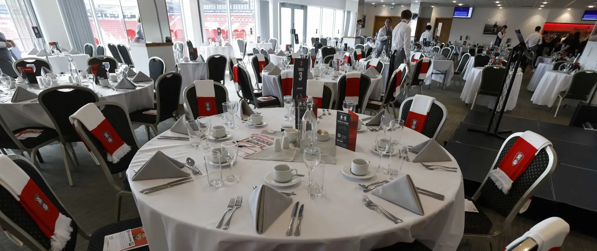 Round banquet tables set in a bright dining hall with white tablecloths, folded napkins, and red chairs.