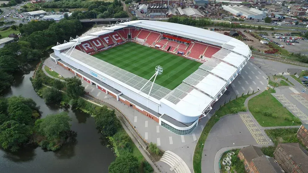 Aerial view of a red-and-white soccer stadium with a green field, surrounded by roads and trees.