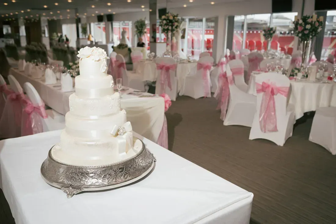 White tiered wedding cake on a silver stand in a decorated reception hall with pink chair sashes.